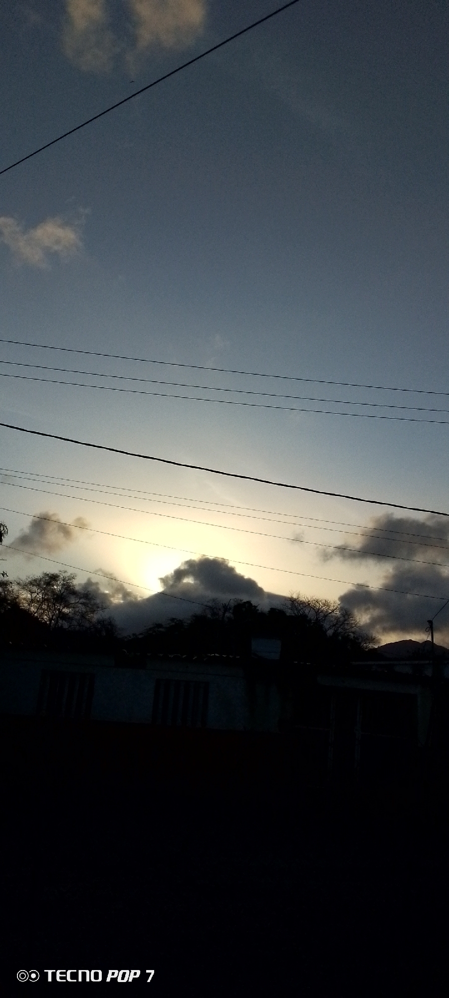 The photograph depicts a scene in Altagracia, Venezuela, taken during twilight or early morning. The sky is a gradient of light blue to pale yellow near the horizon, with scattered clouds. Power lines run across the upper portion of the frame. Silhouetted structures, possibly houses, are visible in the lower half of the image, with dark trees behind them. The lower portion of the frame is almost entirely black due to underexposure, obscuring ground details. The "TECNO POP 7" watermark is visible in the lower left corner.