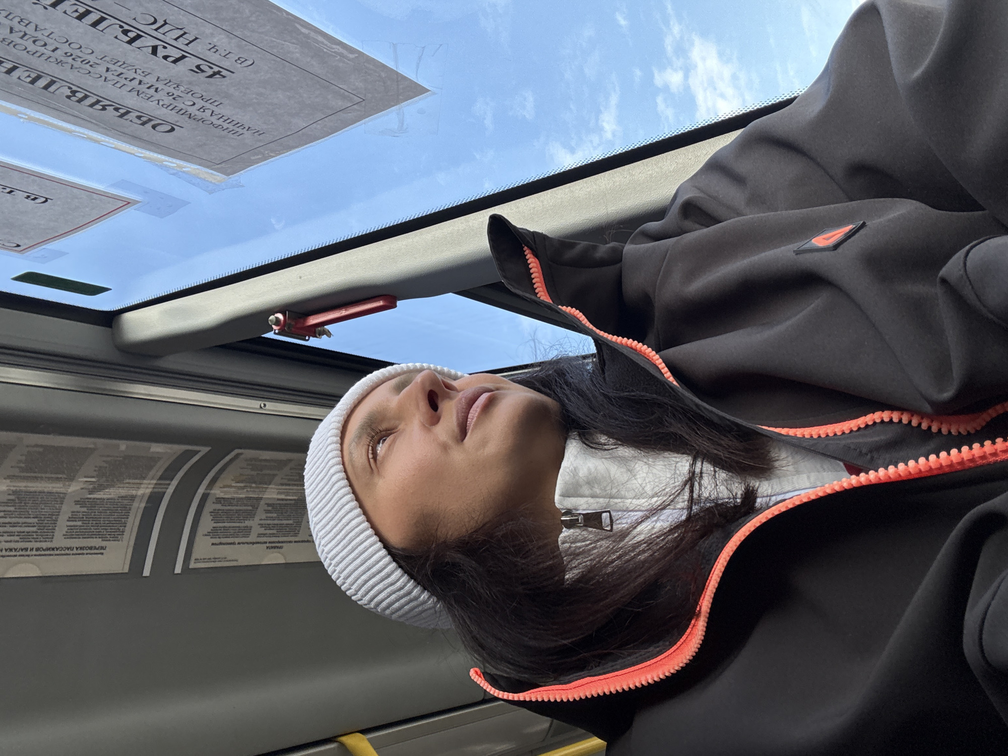 An eye-level indoor shot captures a person looking upwards, likely through a vehicle window or sunroof, in Chaikovsky, Russia. The individual, appearing to be a young woman with long dark hair, wears a white knit beanie and a dark jacket with a prominent coral-orange zipper. Underneath, a white or light-colored garment with a zippered collar is visible. Her eyes are directed towards the sky, and her expression seems calm and contemplative.
The setting is the interior of what appears to be public transport, such as a bus, indicated by the yellow handrail/bar in the lower left and various notices posted overhead. Through the top window, a bright blue sky with scattered white clouds is visible, suggesting clear daytime weather.
Several notices with Russian text are affixed to the vehicle's interior. One prominent notice, partially visible, is titled "ОБЪЯВЛЕНИЕ" (Announcement). It states: "ИНФОРМИРУЕМ ПАССАЖИРОВ" (We inform passengers), "НАЧИНАЯ С 26 МАРТА 2026 ГОДА" (Starting from March 26, 2026), "ПРОЕЗДА БУДЕТ СОСТАВЛЯ" (The fare will be), "45 РУБЛЕЙ" (45 Rubles), and "(В Т.Ч. НДС -" (Including VAT -). Other visible text fragments include "ПЕРЕВОЗКА ПАССАЖИРОВ И БАГАЖА Н" (Transportation of passengers and luggage) and "ПРАВИЛА транспортом" (Rules by transport). The overall scene depicts a moment of quiet transit.