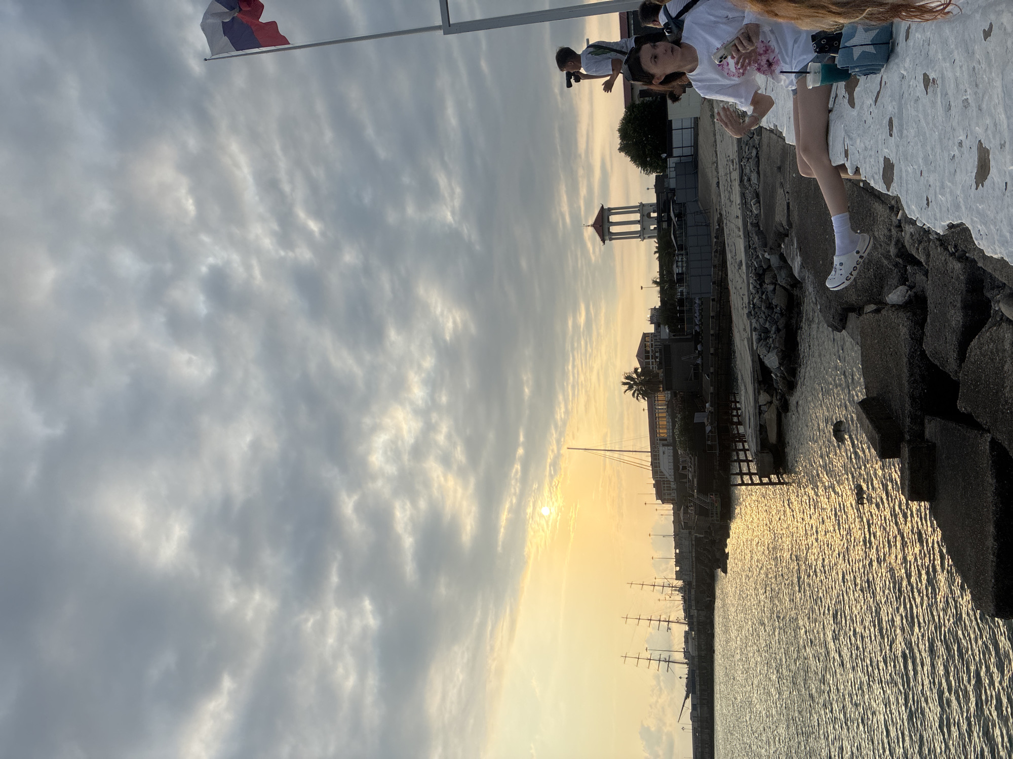 This image depicts a scene at the Sochi Sea Port in Russia during what appears to be sunset. The sun is low in the sky, casting a warm, golden glow on the water and the clouds. Several large sailing ships are docked in the harbor, their masts silhouetted against the bright sky. In the background, buildings and a distinctive white tower with a red roof can be seen along the waterfront.
In the foreground, a young woman is sitting on a low white wall, facing right. She is wearing a white t-shirt with a floral design and denim shorts. She has white socks and white Croc-style shoes. She is holding a smartphone in her left hand and appears to be gesturing with her right hand. Next to her on the wall are a blue drink in a clear cup and a folded blue and white towel. To the right of the frame, another person's shoulder and hair are visible.
Further along the wall and slightly behind the woman, a young person is standing and looking through binoculars. They are wearing a white t-shirt. Behind them, a Russian flag flies from a tall flagpole. The sea is calm, with ripples reflecting the sunlight. The shore is lined with rocks and concrete structures. The overall atmosphere is serene and picturesque, capturing a moment at the end of the day by the sea.