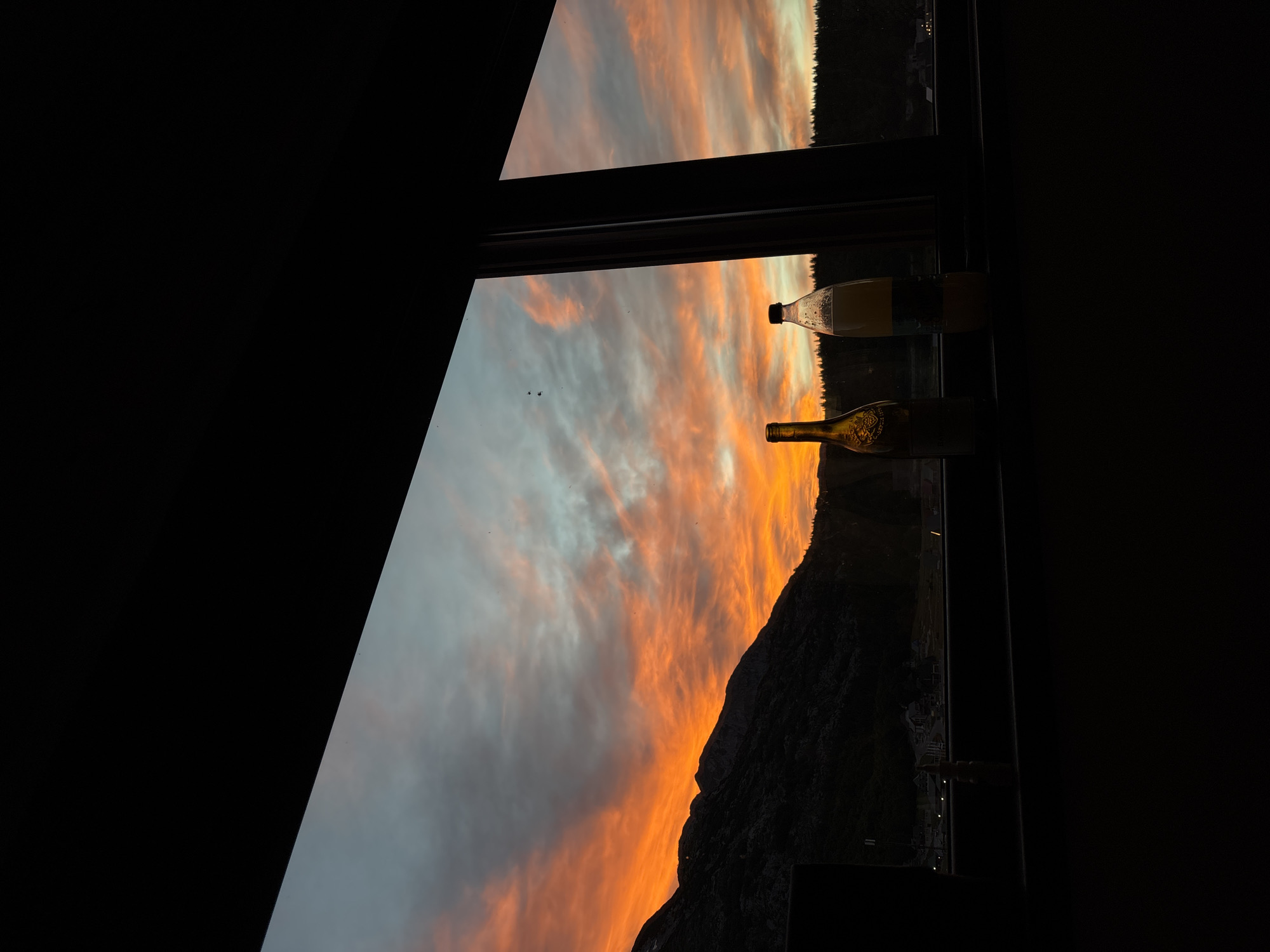 This image depicts a dramatic sunset viewed through a window. The sky is ablaze with vibrant orange and pink hues, with clouds streaked across the canvas. A silhouetted mountain range with trees along its base forms the backdrop. In the foreground, two bottles sit on a windowsill, one appearing to be a wine bottle and the other a clear bottle with a yellow liquid. The scene is framed by dark, silhouetted window frames, creating a sense of looking out from an interior space. There is no visible indication of the location being the city of Virak, Montenegro.