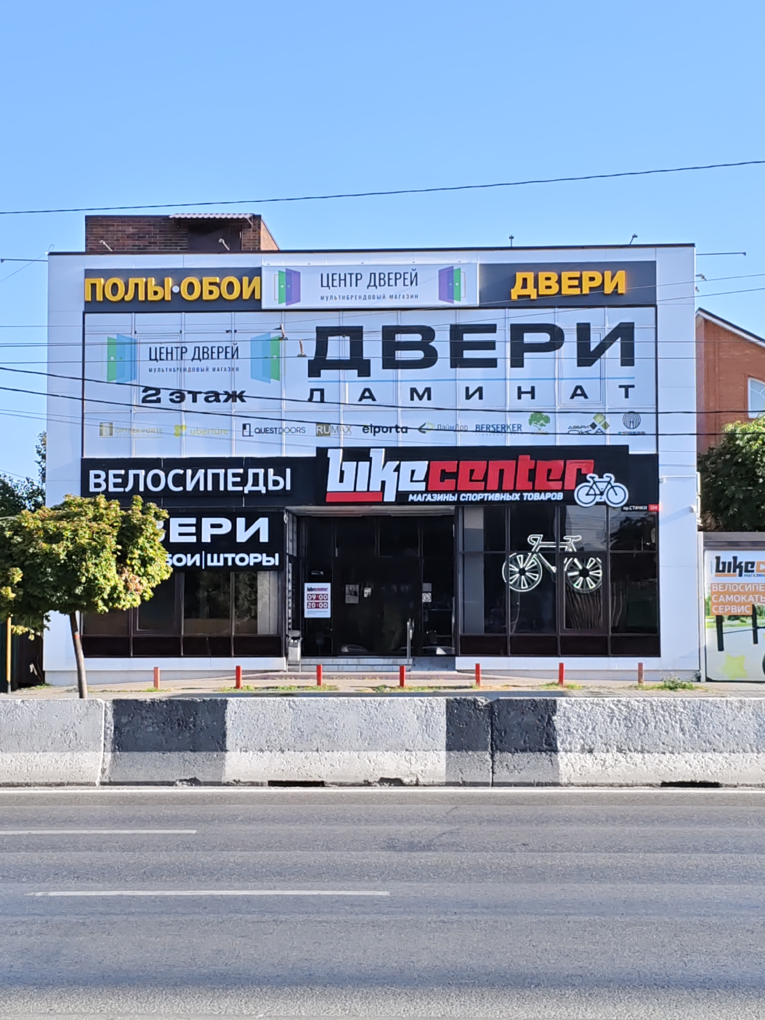 The image shows a storefront in Rostov-on-Don, Russia, featuring multiple businesses. The building has a white facade with prominent signage. On the upper part of the building, the words "ПОЛЫ•ОБОИ" (Floors•Wallpaper) are displayed in yellow lettering. Next to it, "ЦЕНТР ДВЕРЕЙ" (Center of Doors) is written in blue, with the subtitle "МУЛЬТИБРЕНДОВЫЙ МАГАЗИН" (Multi-brand store). To the right, "ДВЕРИ" (Doors) is prominently displayed in large yellow letters. Below this, the word "ДВЕРИ" is written in large black letters with the letters "М И Н А Т" below it, possibly indicating a brand or a specific offering.
Further down, "ВЕЛОСИПЕДЫ" (Bicycles) is written in white letters on a black background. To the right of this, the logo "bikecenter" is displayed in red and white lettering, with the subtitle "МАГАЗИНЫ СПОРТИВНЫХ ТОВАРОВ" (Sports goods stores). A silhouette of a bicycle is also part of the bikecenter logo. The storefront windows display neon bicycle outlines and operating hours from 09:00 to 20:00.
To the far right, a partial sign for "bike C" is visible, with "ВЕЛОСИПЕДЫ" and "САМОКАТЫ" (Scooters) and "СЕРВИС" (Service) also partially visible.
The building is situated in front of a street with a concrete barrier. A tree with green foliage stands to the left of the entrance. The sky is clear and blue. The architectural style appears to be modern commercial.