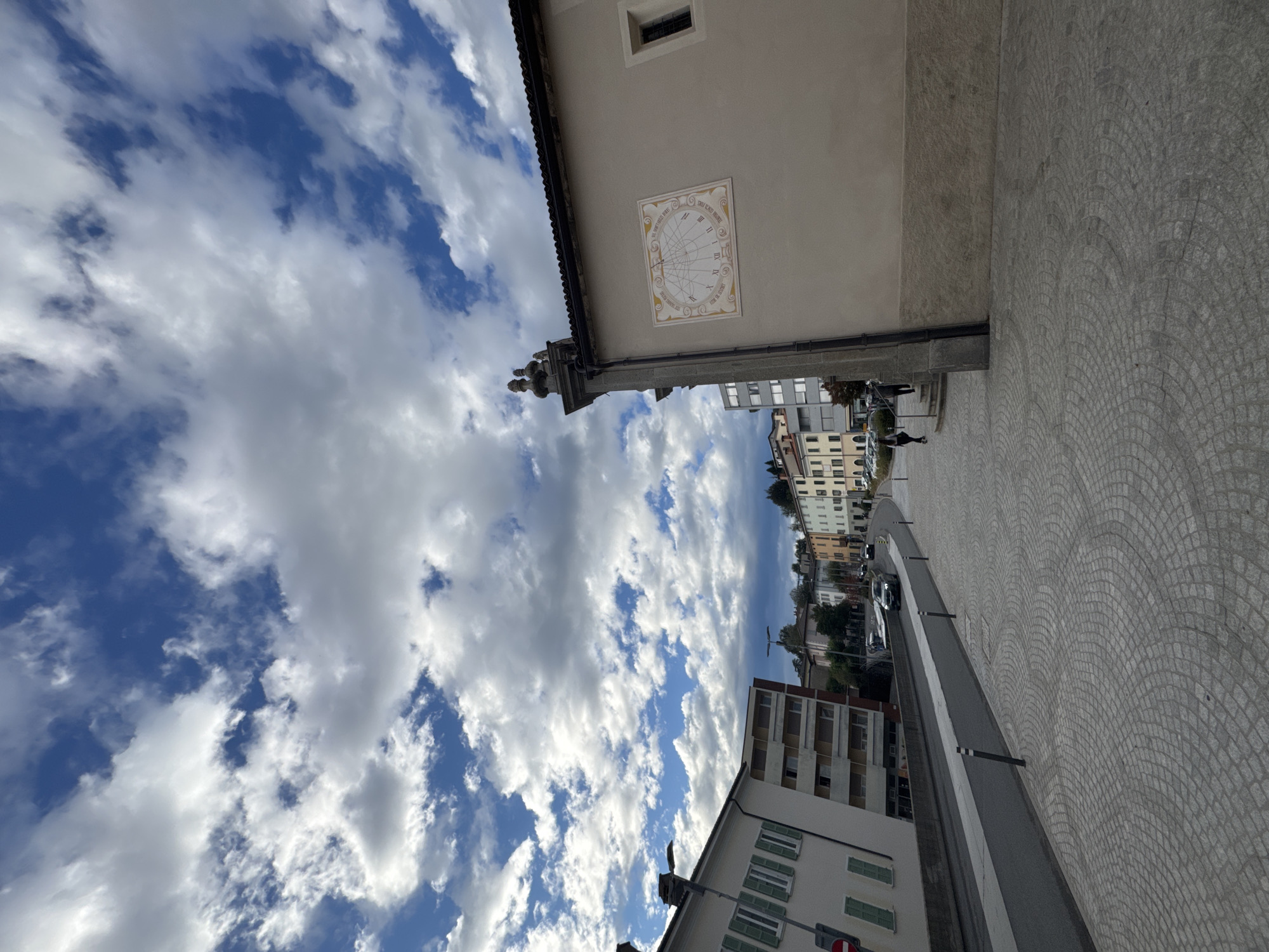This is an outdoor, eye-level, wide shot photograph taken from a pedestrian walkway in the town of Balerna, Switzerland. The walkway is paved with light grey, circular cobblestones and is angled from the foreground left to the right mid-ground. A street with two lanes of asphalt runs parallel to the walkway on the left. Several cars are driving on the street, and streetlights are visible. Buildings line the street on the right side of the image, with the closest building being a light beige structure with a decorative sundial mounted on its exterior wall. The sundial has Roman numerals and intricate lines. Further down the street, a row of multi-story buildings with colorful facades are visible. The sky above is a bright blue, filled with a dramatic display of white and grey cumulus clouds. The overall scene depicts a sunny day in a European town.