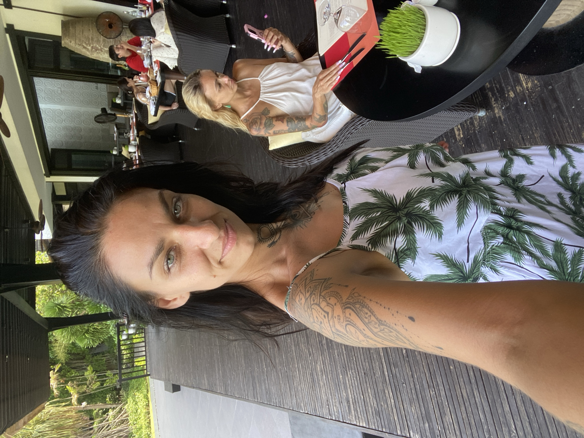 A woman with dark hair and tattoos on her arm and neck is taking a selfie at an outdoor restaurant. She is wearing a white dress with a palm tree print. Behind her, a blonde woman is looking at her phone at a table with a menu and a glass. In the background, other people are seated at tables, and there is lush greenery visible. The flooring is made of dark wood planks.