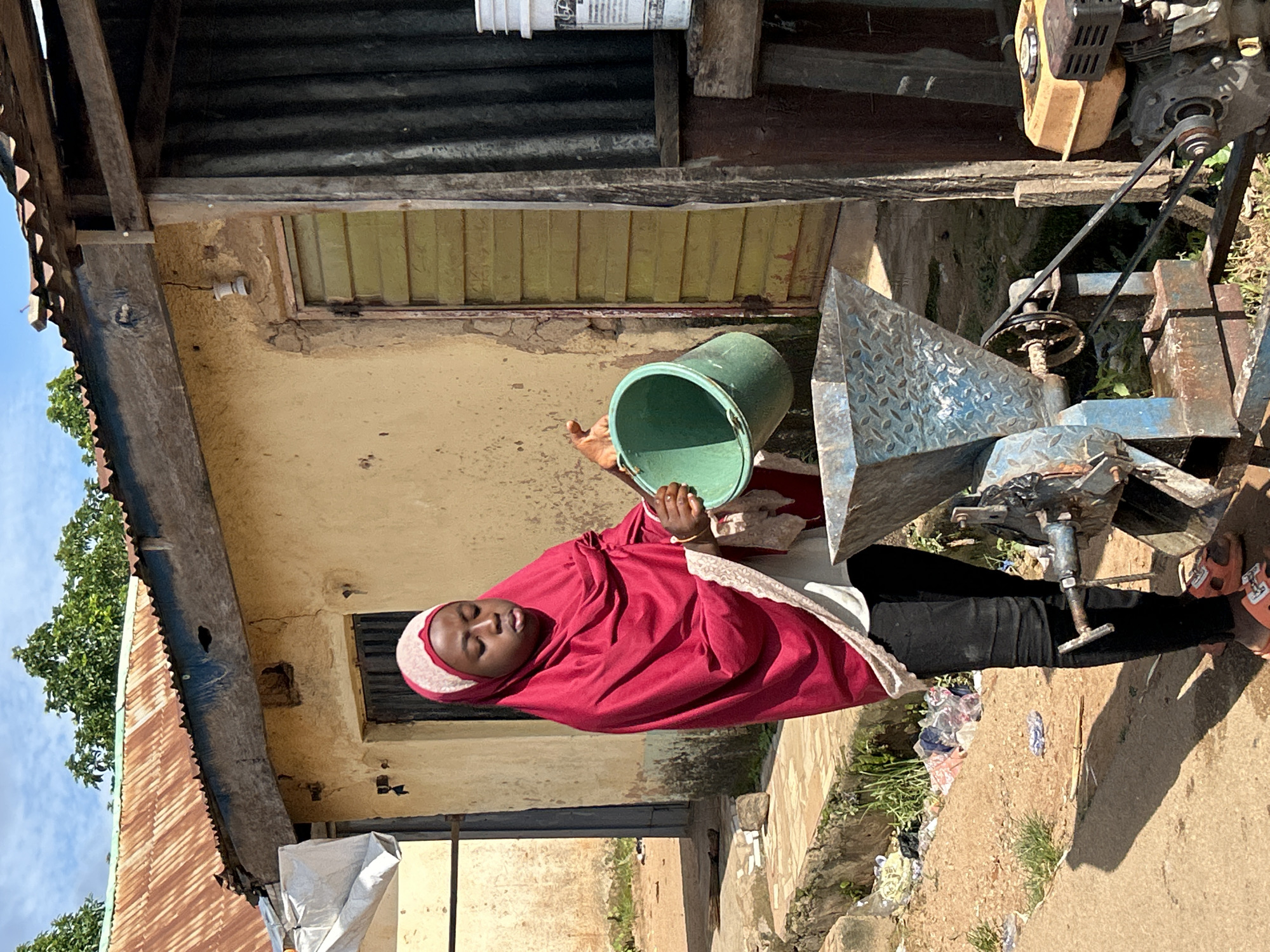 In the city of Jos, Nigeria, a young person wearing a red headscarf and black pants stands outdoors, pouring from a green bucket into a metal grinding machine. The machine, which appears to be powered by a small yellow engine, is positioned on a dirt ground. Behind the person and machine, a weathered yellow building with a corrugated metal roof is visible. The building has a green-painted door and a broken light fixture hanging from the eaves. To the right, a portion of a darker corrugated metal structure is present. The environment is dusty and somewhat unkempt, with debris scattered on the ground. The sky above is partly cloudy.