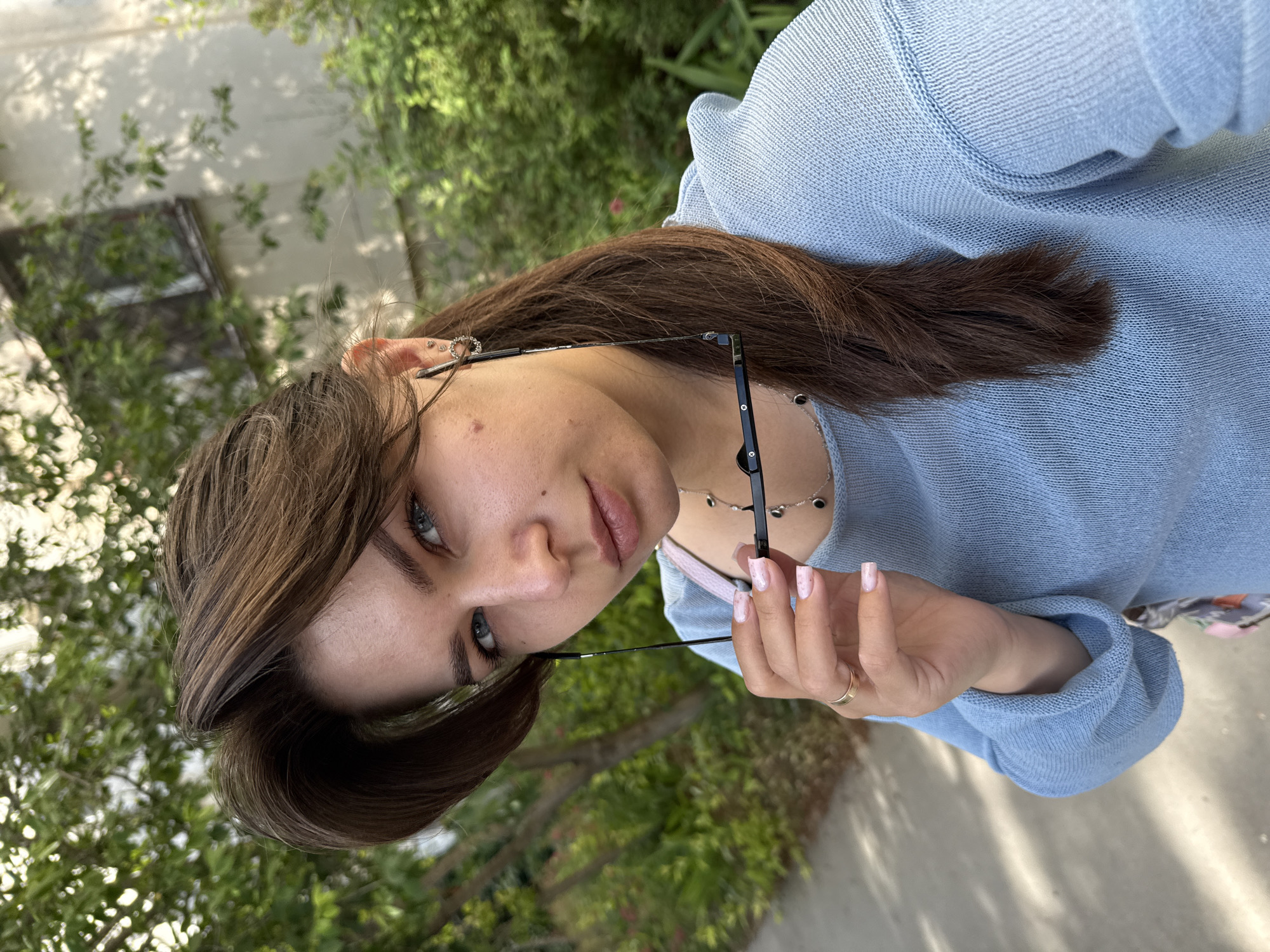 A young woman with medium-length brown hair and blue eyes is taking a selfie outdoors. She is wearing a light blue, long-sleeved sweater and is holding a pair of glasses in her right hand. She has a gold ring on her left ring finger and is wearing a necklace with several small black circular pendants. She has on stud earrings with a circular design. The background shows green trees, a building with windows, and a sidewalk. The lighting suggests it is daytime.