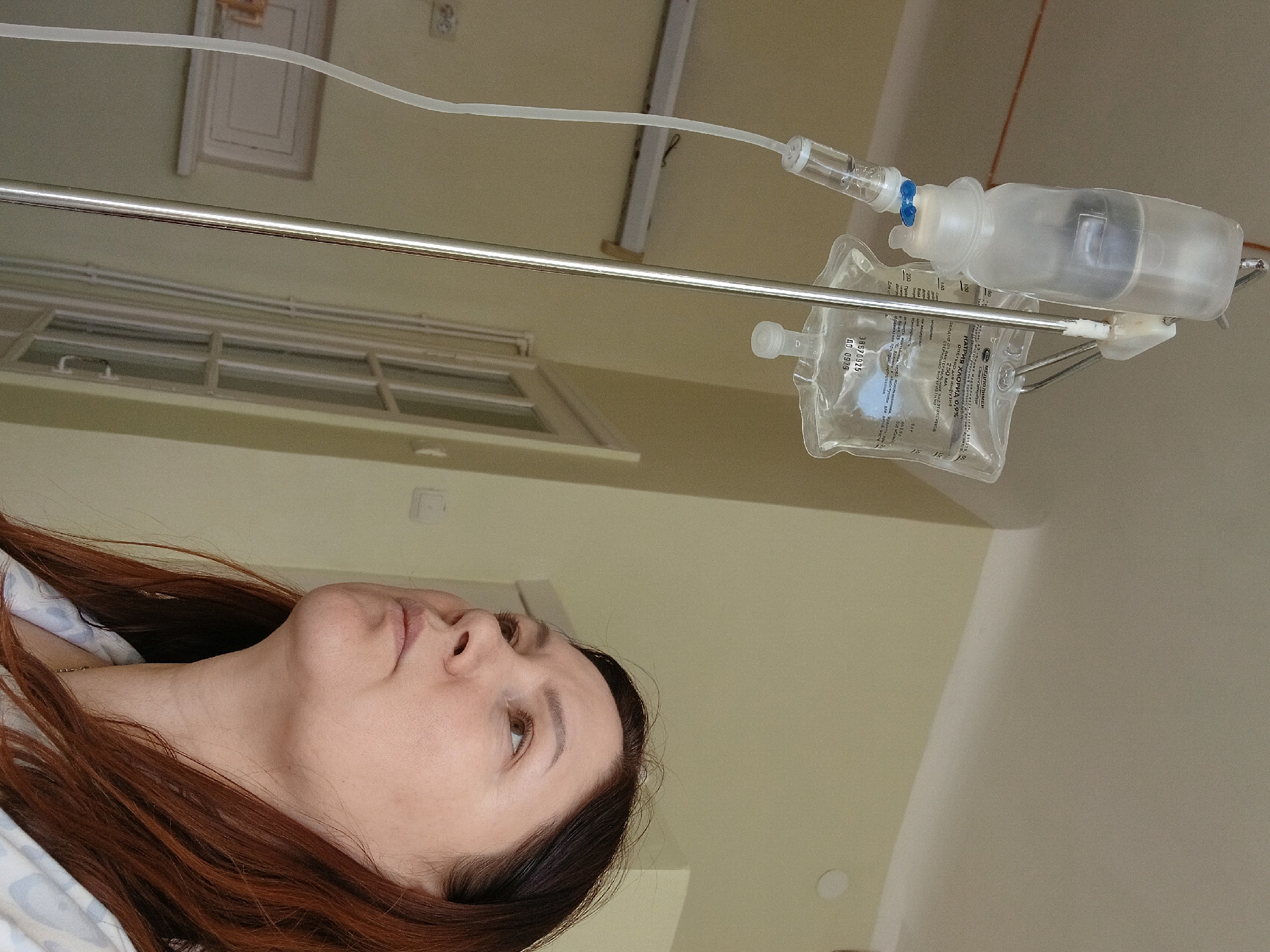 The image shows a woman lying in a hospital bed, viewed from a low angle. An IV bag and equipment hang above her, attached to a metal pole. The IV bag has a label in Russian indicating it contains sodium chloride 0.9% and is produced by "МЕДПОЛИМЕР" (Medpolymer) in Saint Petersburg, Russia. A window is visible in the background, and the room appears to be a standard hospital room with neutral-colored walls.