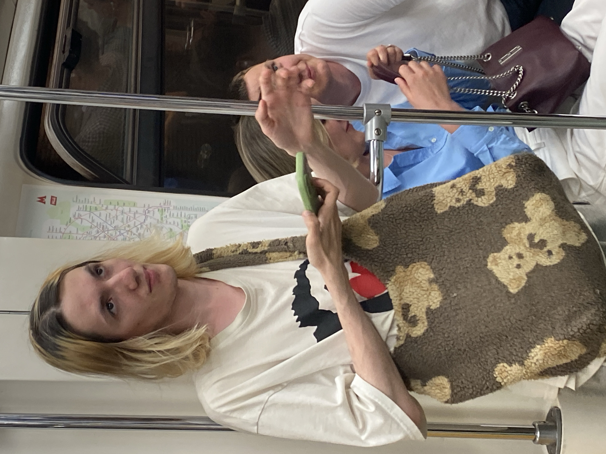 In Moscow, Russia, inside a subway car, a young person with shoulder-length blonde hair, parted in the middle, stands holding a large, brown fuzzy tote bag adorned with teddy bear motifs. They are wearing a light-colored t-shirt with a graphic design. To their right, a person in a light blue collared shirt is partially visible, and further to the right, a man in a white t-shirt is looking to his left. The man is also holding a dark red purse with a silver chain. The environment includes a vertical metal pole and a subway map on the wall.