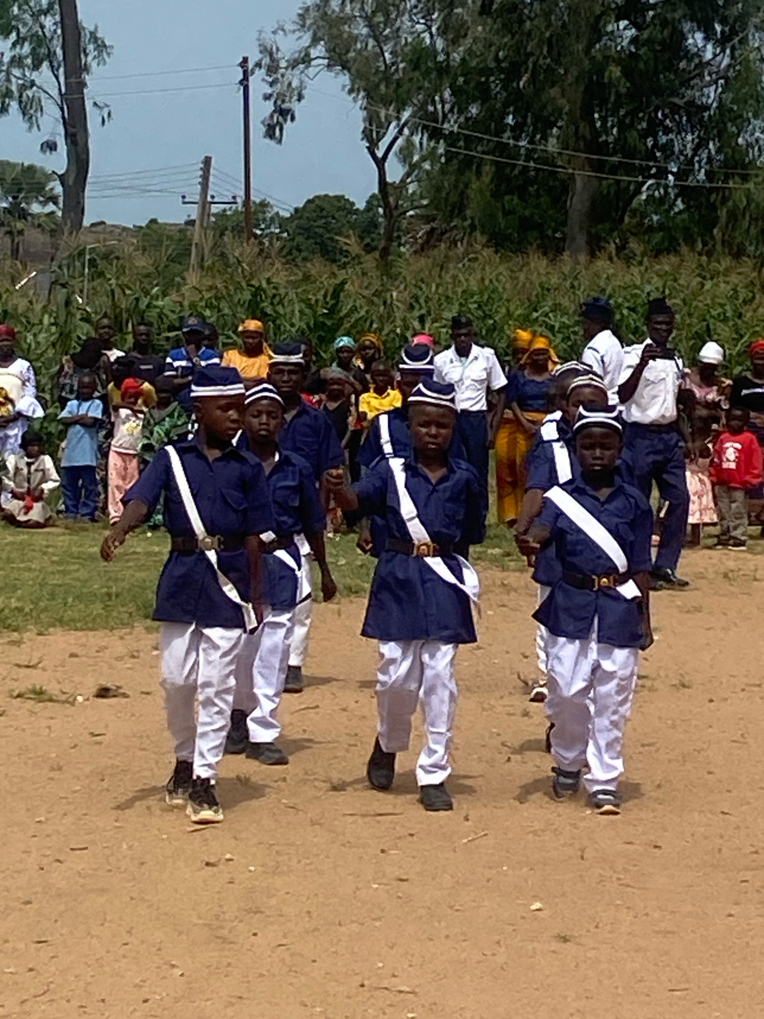 The image depicts an outdoor event in Binchi, Nigeria, where a group of children in uniform are marching. The children are wearing dark blue jackets with white sashes and white pants, and matching caps. They appear to be participating in a parade or ceremony. In the background, a crowd of adults and other children are gathered, some watching the marching group. The environment is predominantly dirt ground, with trees and vegetation visible in the distance, suggesting a semi-rural or open area within the city. The sky is overcast.