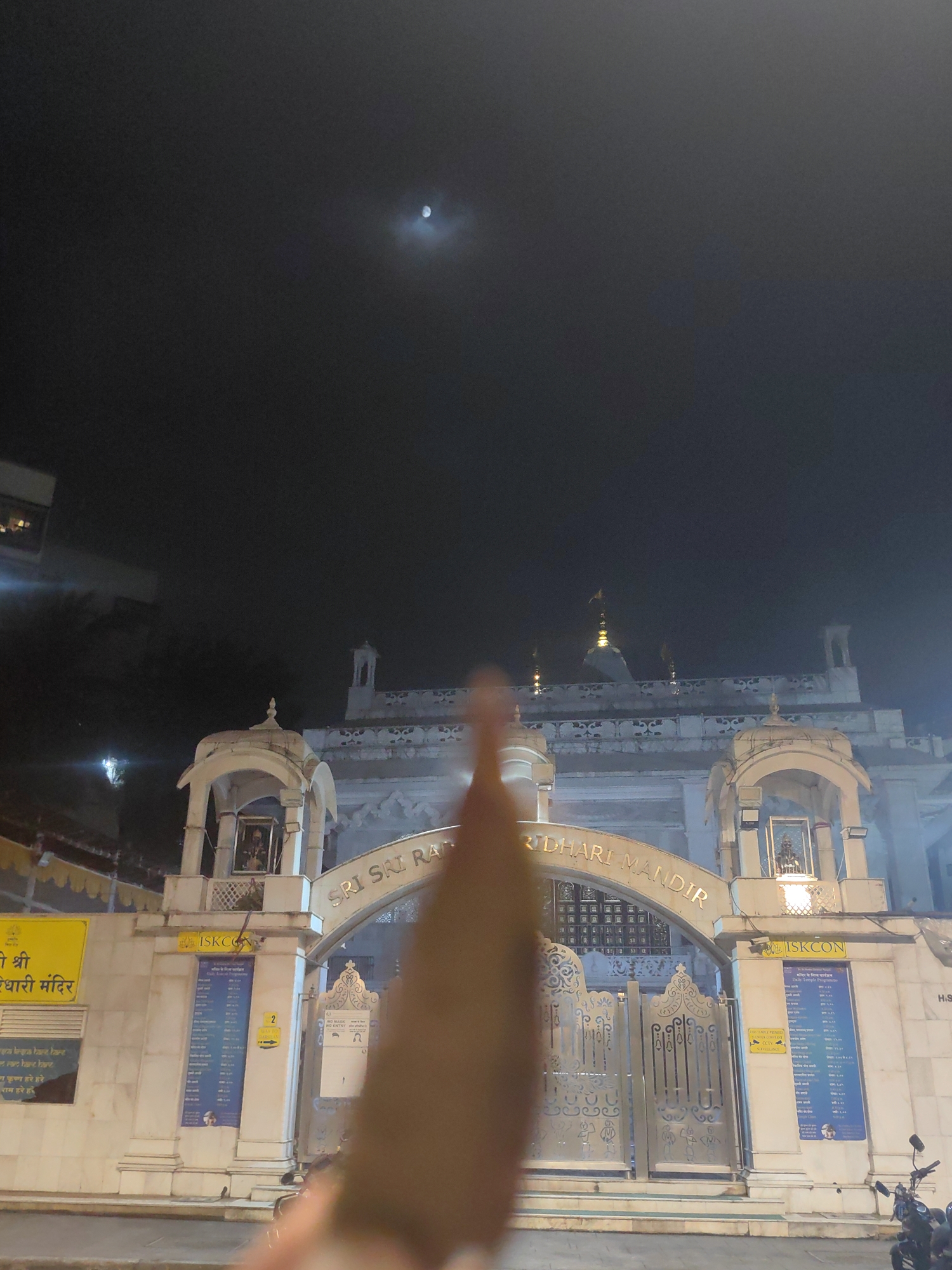 The image shows the entrance to the ISKCON Radha Rasbihari Temple in Mira Road, Mira-Bhayander, India, at night. The temple's white facade is illuminated, with ornate gates and arched entrances. A sign above the main gate reads "SRI SRI RADHA RASBIHARI MANDIR." A smaller yellow sign on the left displays text in Hindi. Several informational posters are affixed to the wall to the right of the entrance. A portion of a person's hand and thumb is visible in the foreground, obscuring part of the scene. The sky above is dark with a faint moon visible. A motorcycle is parked near the entrance.