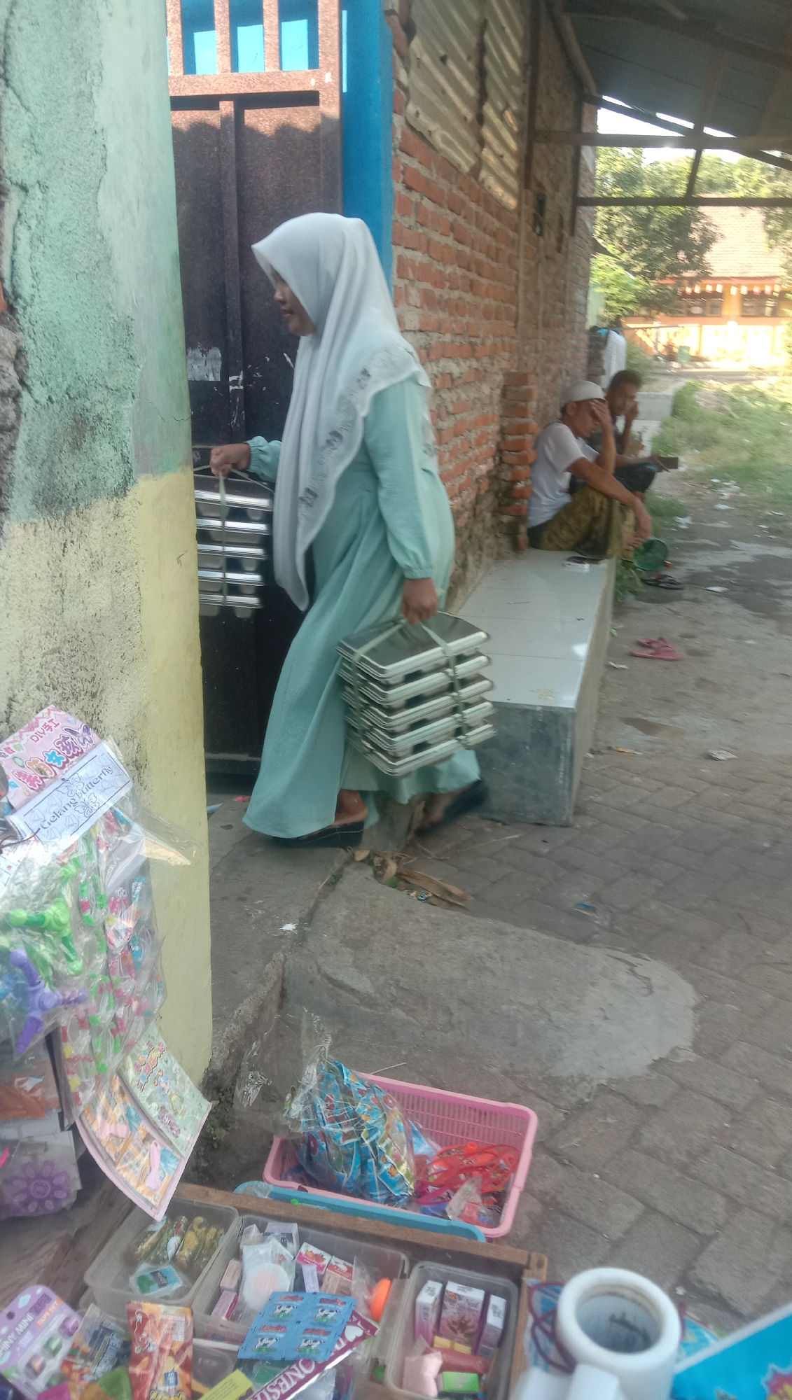 A woman wearing a light blue hijab and dress walks to the right, carrying a stack of metal trays in each hand. She is outdoors next to a building with a green and yellow textured wall on the left and a brick wall on the right. Two men are seated on a concrete bench in front of the brick building, with the man on the left wearing a white cap and the man on the right wearing a dark cap. In the foreground to the left, there are several clear plastic containers filled with small items, possibly toys or snacks, and a pink basket containing more items. The ground is paved with stones, and there are some loose branches and debris scattered around. The scene appears to be in a marketplace or street in Karang Anyar, Indonesia.