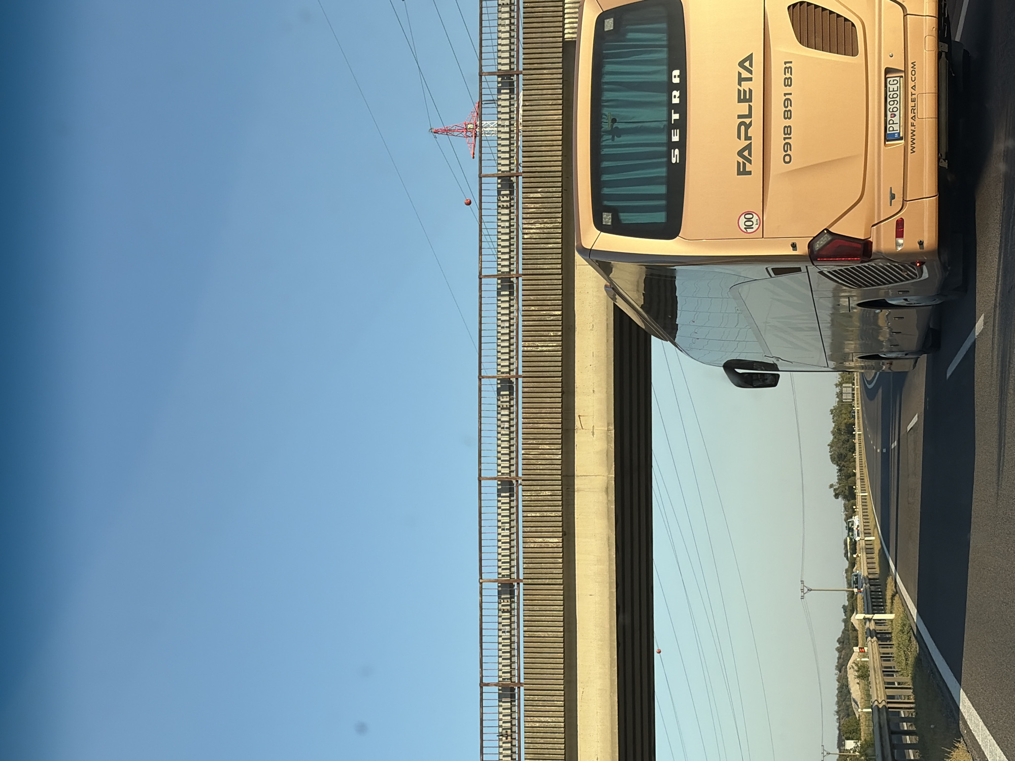 The image shows the rear of a gold-colored Setra bus on a highway, likely near Sereď, Slovakia, given the license plate. The bus has "FARLETA" written on its back, along with a phone number "0918 891 831" and a website "WWW.FARLETA.COM". A speed limit sticker of "100 km" is also visible. The bus is in motion, as indicated by the motion blur and the perspective of the road. The license plate is Slovakian, reading "PP 696EG". The bus is on a multi-lane highway with a concrete overpass above. In the background, there are power lines, some vegetation, and a red radio tower. The sky is clear and blue.