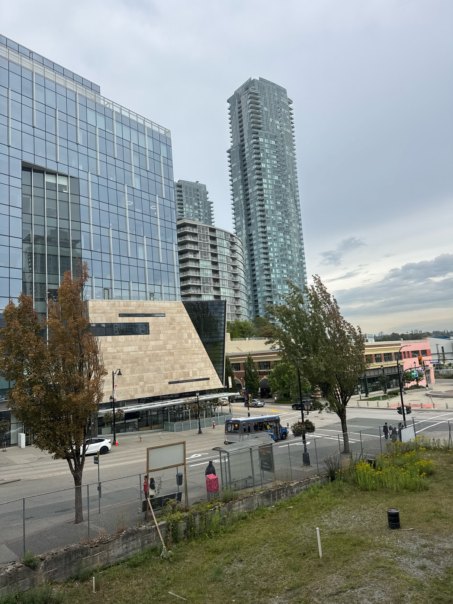 The image depicts a street scene in downtown New Westminster, Canada, on an overcast day. In the foreground, a chain-link fence runs along the edge of a grassy embankment. Beyond the fence, a sidewalk runs parallel to a street. Several trees, some with autumn foliage, are planted along the sidewalk.
Dominating the left side of the frame is a modern glass and stone office building with a distinctive angled facade. To the right of this building, a tall, slender residential skyscraper with numerous balconies stands out against the sky. Further in the background, other buildings, including a pink commercial structure, are visible.
On the street, traffic is moving. A blue transit bus is stopped at a bus shelter, where a few people are visible waiting. A white car is parked on the street. Pedestrians are seen on the sidewalk. The environment appears urban, with a mix of commercial and residential architecture.
