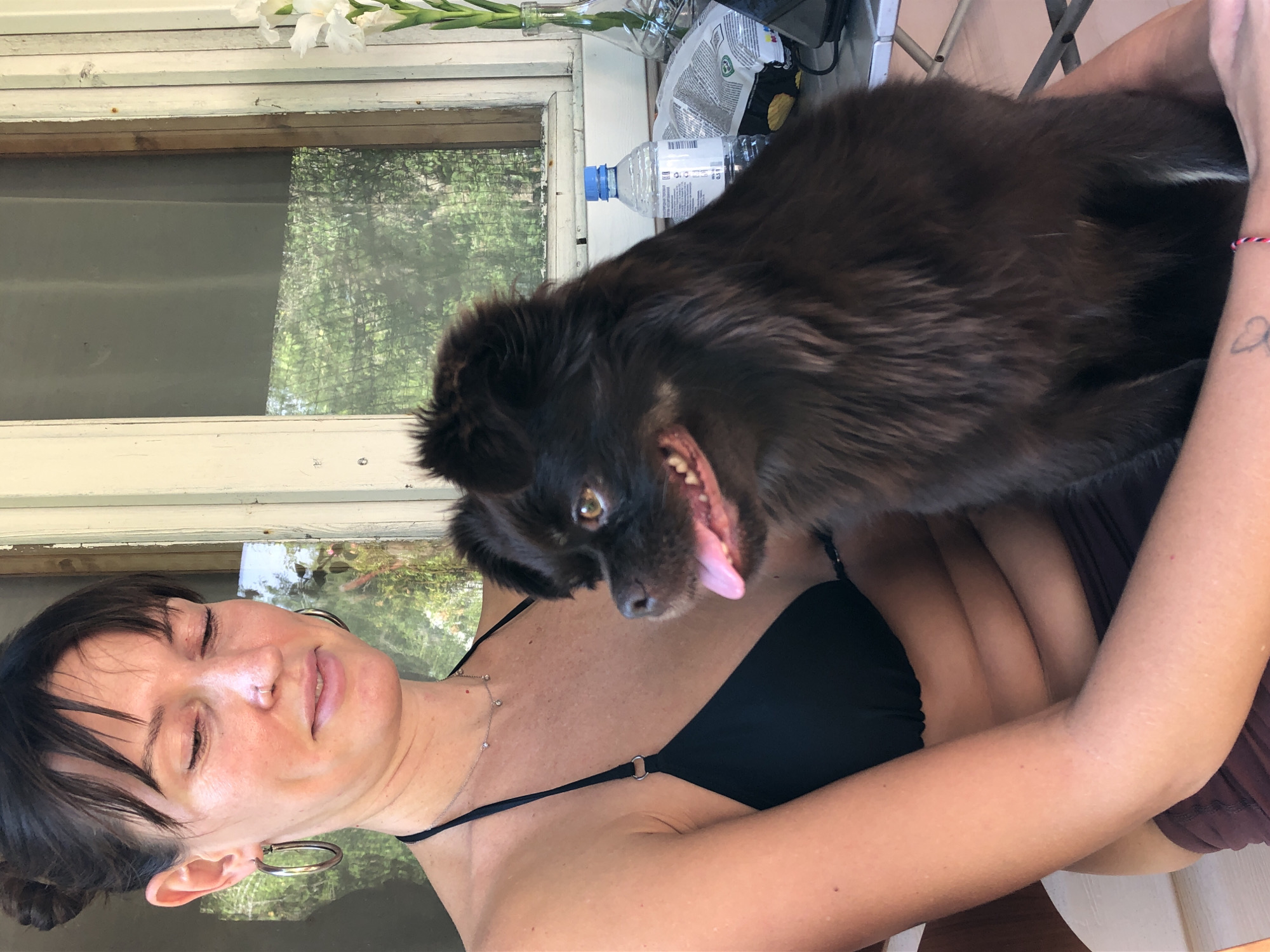 A medium shot captures a woman in a black bikini top holding a dark brown, fluffy dog. The woman has dark hair pulled back in a bun and is looking down and to her left, with her eyes partially closed and a slight smile. She wears hoop earrings and a delicate necklace. The dog, positioned on her lap and right side, looks towards the camera with its mouth slightly open and tongue visible, appearing to pant or smile. The background shows a rustic wooden structure, possibly a porch or balcony, with a window covered by a screen through which trees and foliage are visible. A glass vase with white flowers, a bottle of water, and a bag of chips are on a table in the background. The setting appears to be outdoors, possibly a dacha or a rural residence. There is no discernible information in the image that can specifically pinpoint the location to Красная Пресня Снт, Russia.