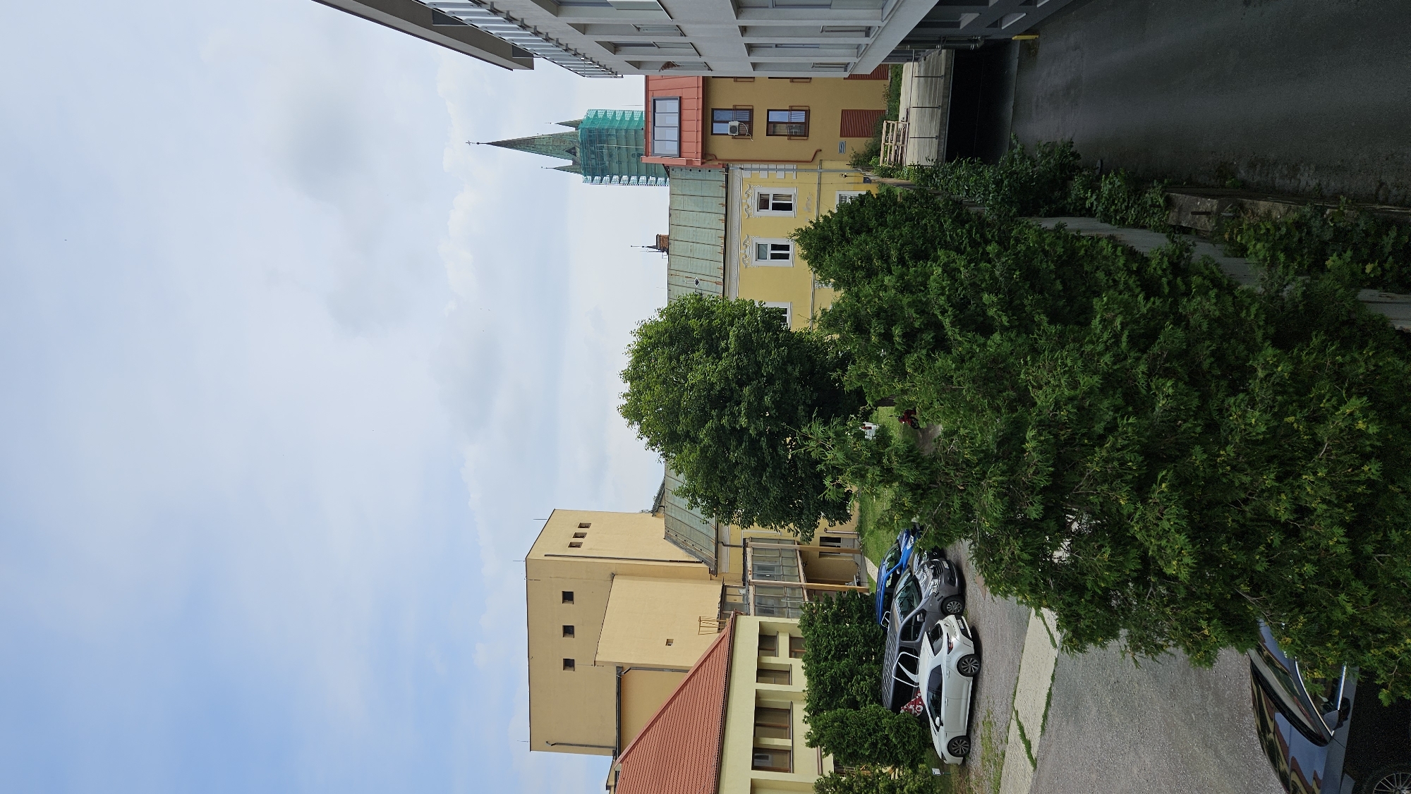 The image captures a view of an urban area, likely in Prešov, Slovakia, featuring a mix of architectural styles and green spaces under a cloudy sky. In the foreground, several evergreen trees and bushes line a concrete wall, with a dark colored car partially visible. A parking lot with several cars, including a white hatchback and a gray minivan, is located to the left.
Behind the trees, several buildings are visible. To the left, a building with a red tiled roof and yellow walls stands next to a larger, taller yellow building with a distinctive boxy shape and small square windows. In the center, a cluster of buildings includes a yellow house with a red-orange roof and a green-roofed structure. A notable feature in the background is a tall green spire, likely part of a church, covered in scaffolding, indicating ongoing renovation or construction. To the far right, a modern building with white facade and multiple windows is partially visible. The overall impression is one of an older European city with pockets of modern development and preserved historical elements.