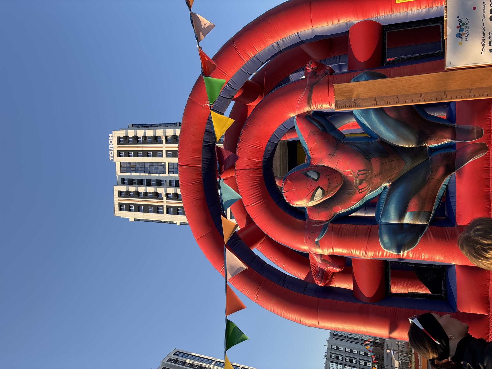The image depicts a Spiderman-themed inflatable play structure, complete with pennant flags, set against a clear blue sky and several modern high-rise buildings. The building on the top center has the word "ТАЛАН" on it. This play structure likely stands in an urban area, based on the high-rise buildings visible in the background. The photograph seems to be taken from ground level, looking up at an angle toward the inflatable structure and the buildings. Some people are visible on the right side of the image. Based on "ТАЛАН", the city of Perm, Russia is likely.