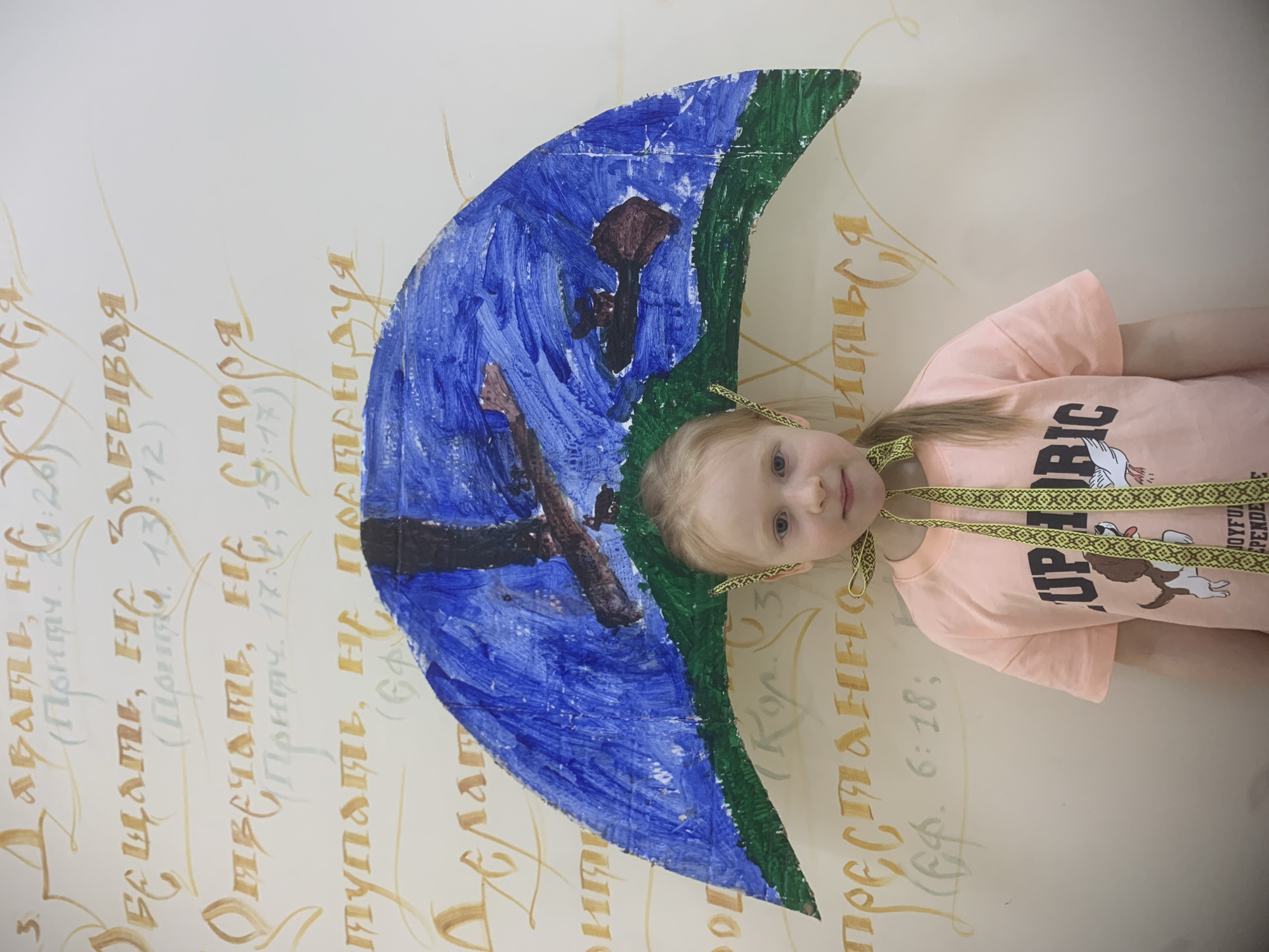 A child wearing a homemade paper moon headpiece, painted blue and green, poses indoors in front of a wall decorated with calligraphy. The calligraphy appears to be in Russian. The child wears a peach-colored t-shirt adorned with a cartoon dog and the word "UPIRIC," along with a patterned ribbon tied around the neck. The location is possibly Izhevsk, Russia, based on the calligraphy.