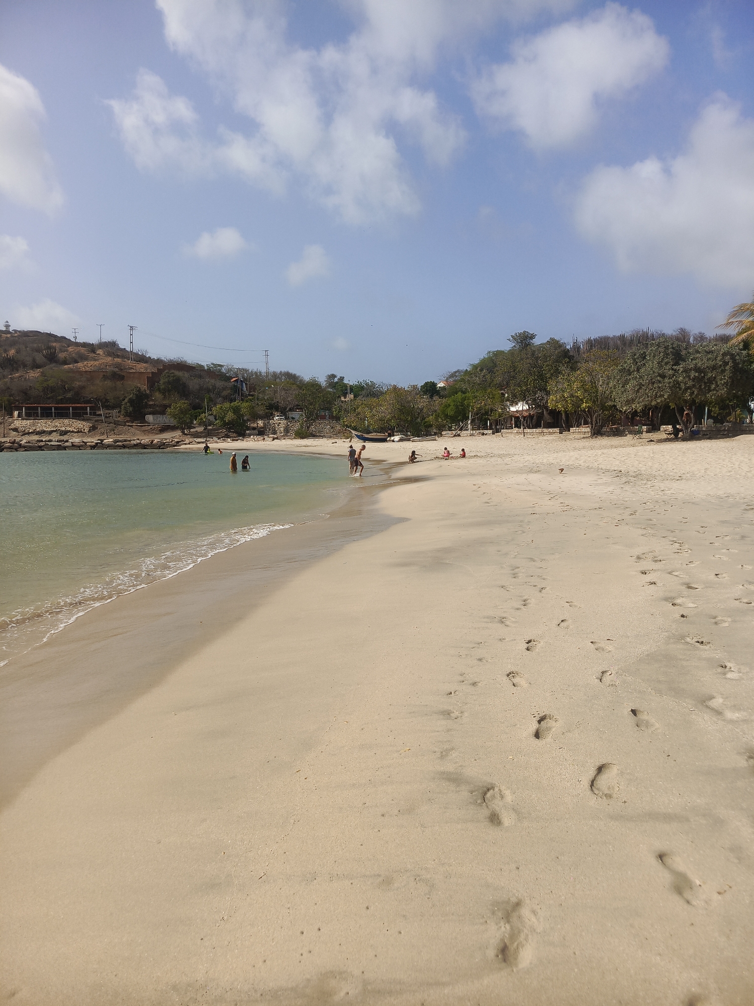 The image shows a beach scene in Pedro González, Venezuela, under a sky with scattered clouds. The beach features light-colored sand with numerous footprints. The shoreline is visible with gentle waves. In the background, there is vegetation and some buildings on a low hill. Several people are present: some are swimming in the water, while others are standing or playing on the beach.