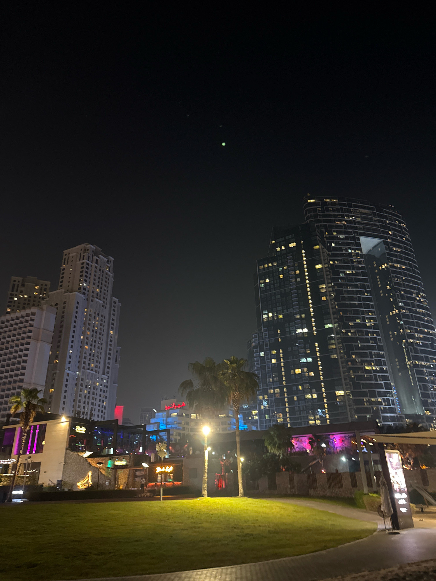 The image depicts a nighttime view of a modern urban landscape in Dubai, United Arab Emirates, likely in the Jumeirah Beach Residence (JBR) area. Dominating the scene are several tall, illuminated skyscrapers, characteristic of Dubai's architecture. On the right, a distinctively curved skyscraper with a large, open section near the top is visible. To the left, a cluster of more rectilinear, high-rise buildings are lit up, showcasing numerous windows. In the foreground, a well-maintained green lawn stretches across the frame, bordered by a paved walkway. Adjacent to the lawn is a low-rise structure with a vibrant, illuminated facade, suggesting it houses restaurants or entertainment venues, with palm trees interspersed throughout the area. Several streetlights are present, casting pools of light on the walkway and lawn. The sky is dark, indicating it is nighttime, with a few faint, indistinct lights or celestial bodies visible. There is a general sense of a lively, developed urban environment.