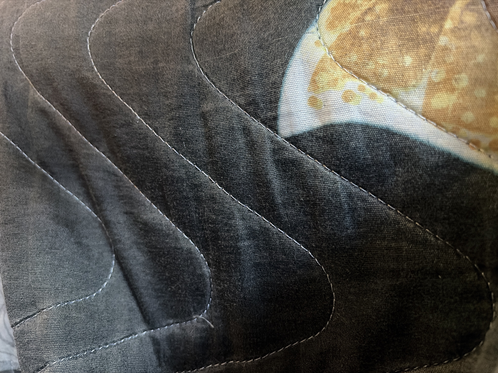 This is a close-up, overhead shot of a quilted fabric. The fabric is dark gray with wavy white stitching. Part of a pattern with yellow and white circles can be seen on the right side of the image. The quilted fabric is likely part of a blanket or a piece of clothing.