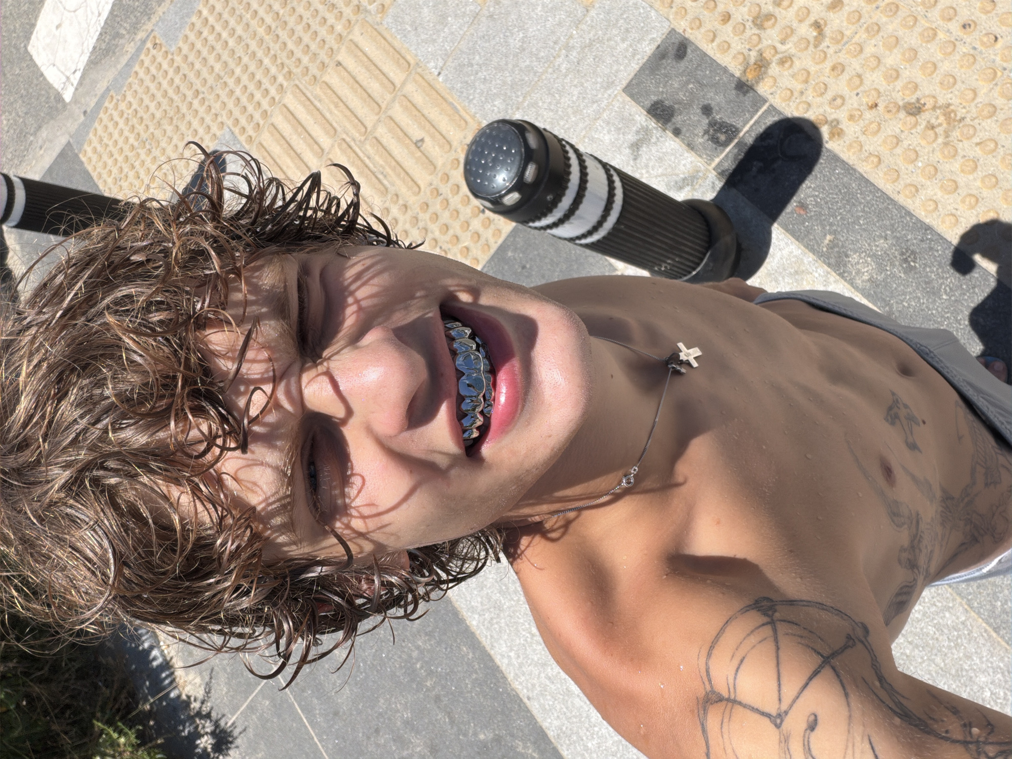 The image is a high-angle, close-up, selfie-style shot of a shirtless young man with wet, curly brown hair and a metallic grill on his teeth. He is wearing a silver necklace with a cross pendant. The man is standing outdoors on a paved surface with a tactile paving pattern. In the background, there is a black and white striped bollard. The lighting suggests it is a sunny day. The image does not contain any specific visual cues to identify a location in Busan, South Korea.
