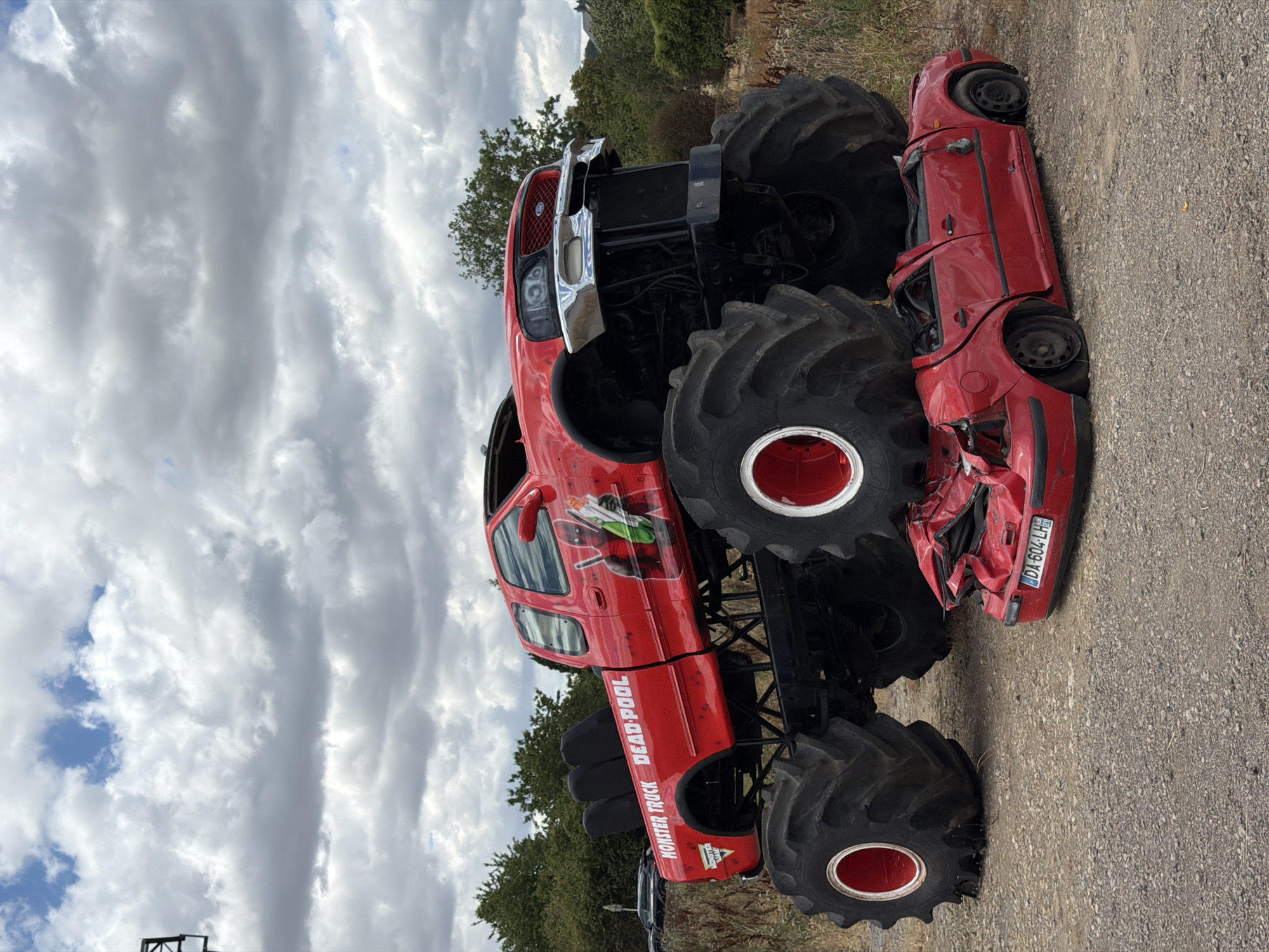 In Esch-sur-Alzette, Luxembourg, a large red monster truck, adorned with a "Deadpool" graphic on its side, is positioned over a crushed red car. The monster truck, featuring oversized tires and a modified chassis, has its front tires resting on the roof and hood of the smaller vehicle, causing significant deformation. The car appears to be a sedan, with its doors open and its body severely damaged, tires deflated. The scene takes place outdoors on a gravel surface, with dry vegetation and trees visible in the background under a cloudy sky. No people are present in the immediate vicinity of the vehicles.