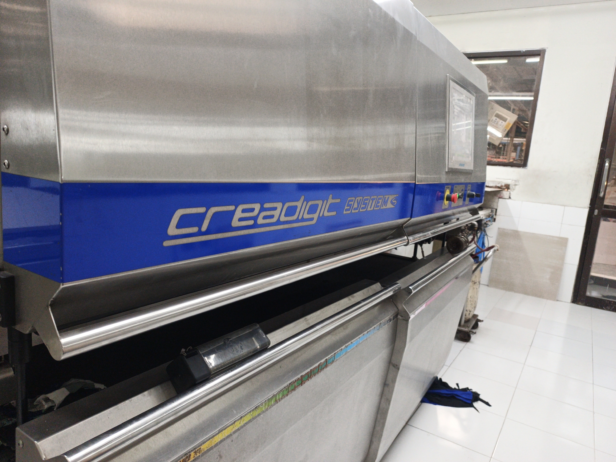 An interior shot captures a large, stainless steel industrial machine with a prominent blue stripe bearing the silver text "creadigit SYSTEMS". The machine is positioned within what appears to be a workshop or factory setting, characterized by white tiled flooring and walls. To the right, a window offers a glimpse of the exterior, showing reflections of industrial structures and possibly a loading bay. The machine itself features metallic rails along its lower section, and a black object, possibly a sensor or part of the machinery, is visible on one of these rails. A partially opened blue and black bag rests on the floor near the base of the machine. The overall impression is one of an operational industrial environment.