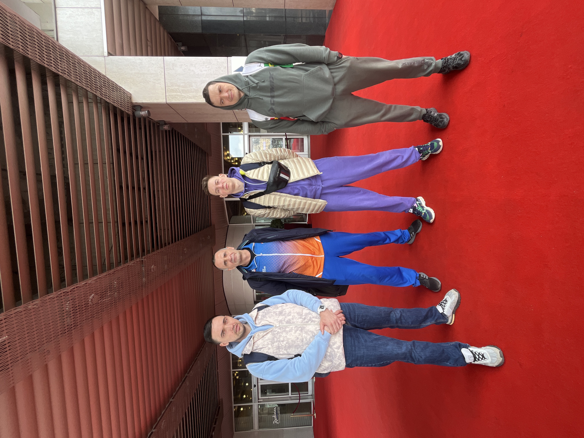 Four men are standing in a line on a red carpet in front of a building. The men are all dressed in casual clothing. The building has a modern design with red metal accents. The Radisson Hotel is visible in the background. The image was taken in Moscow, Russia.