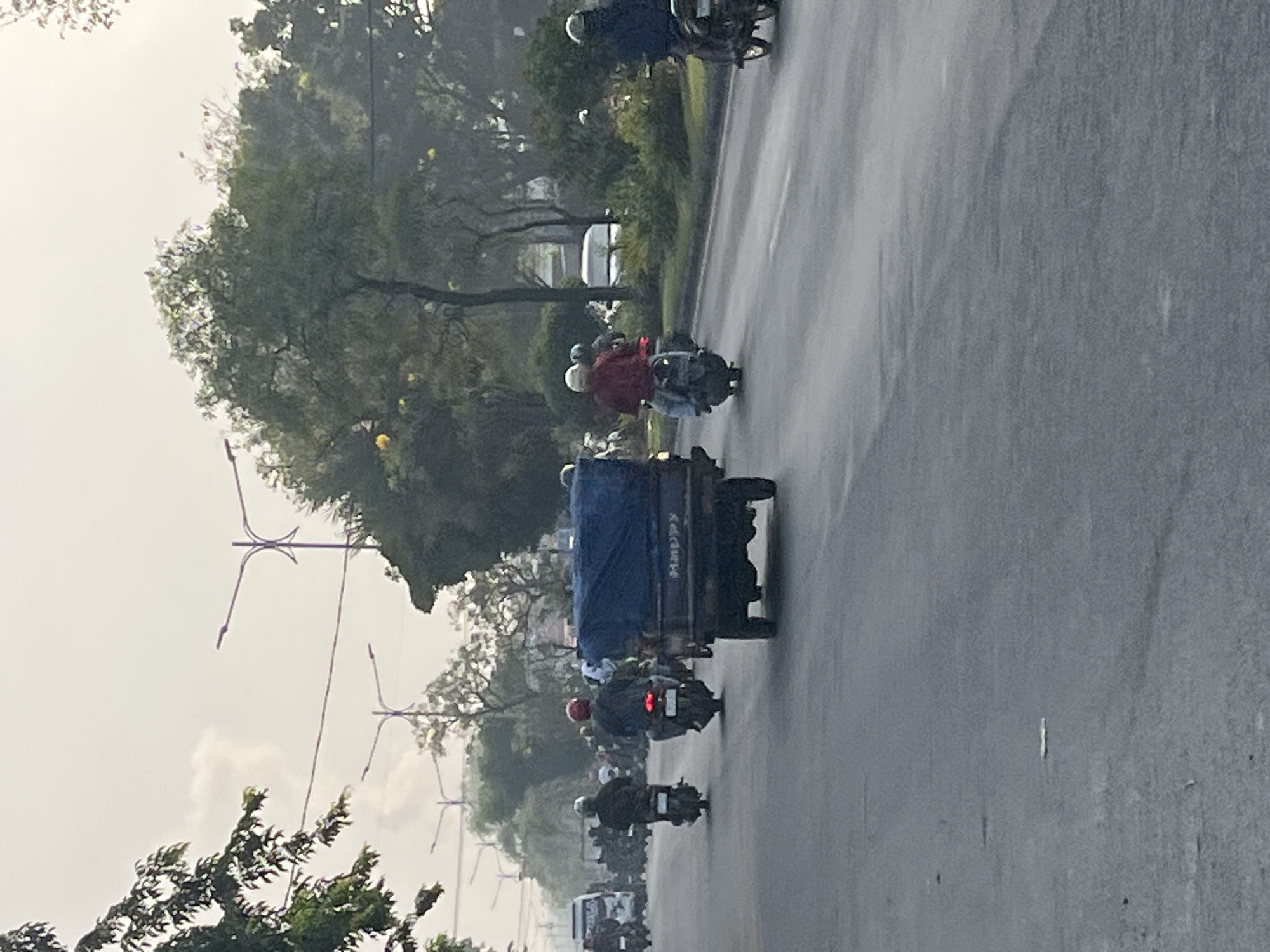 The image depicts a street scene in Gelam, Indonesia, characterized by a moderate level of traffic. In the foreground, a three-wheeled vehicle with a blue tarp covering its cargo is traveling on the right side of the road. Several motorcycles are also visible, with riders wearing helmets. Some motorcycles are in motion, while others appear to be part of a larger group. The road surface is asphalt and appears to be in good condition. The environment includes trees and vegetation lining the sides of the road, with streetlights present, indicating an urban or suburban setting. The sky is overcast, suggesting a cloudy day. The overall impression is that of a typical day in a populated area with active transportation.