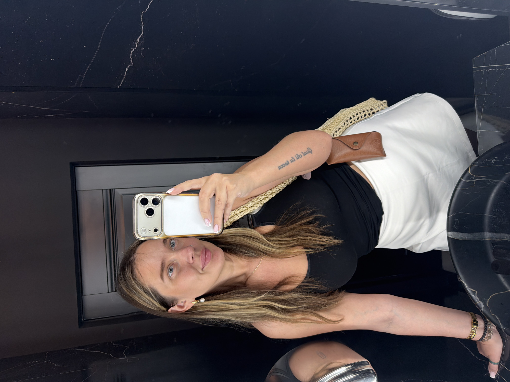 The image shows a young woman with long, blonde-brown hair, wearing a black tank top and a white skirt. She is holding a smartphone and taking a selfie in a bathroom with dark marble walls. She has a gold necklace and earrings, a tattoo on her arm, and a woven bag with a brown glasses case attached. The location is identified as Укромновское сельское поселение, Ukraine, though there are no visual cues in the image to support this specific location. The woman is looking at the camera with a slight smile.