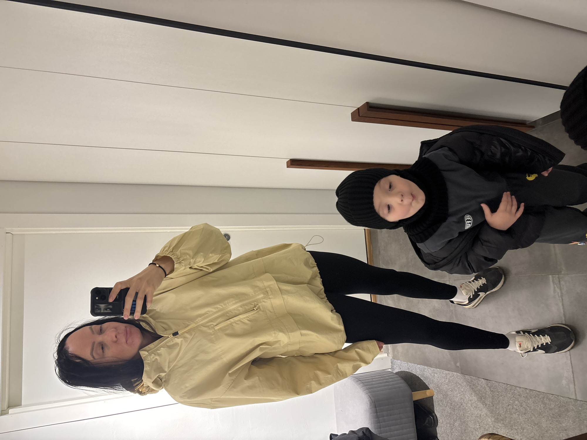 A woman is taking a selfie in a room with a child visible in the reflection of a mirror. The woman is wearing a light yellow jacket and black leggings. The child is wearing a black jacket and a black beanie. The room has a light-colored wall with a dark trim. There are also shelves visible in the mirror reflection. The location is Chaikovsky, Russia.