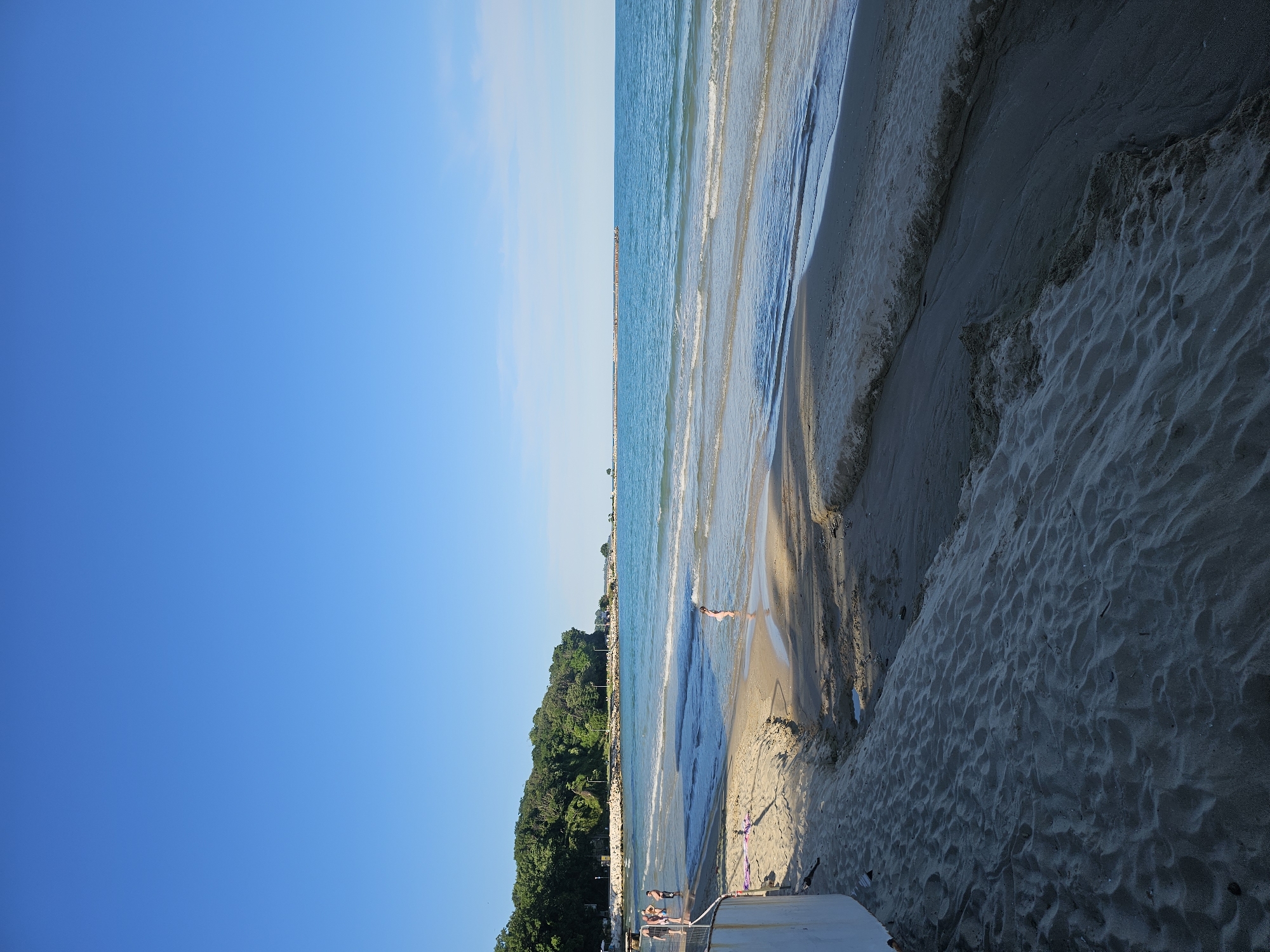 The image depicts a beach scene on a clear, sunny day. The sky is a vibrant, cloudless blue, suggesting fair weather and likely midday or late afternoon. The composition is divided, with the left side dominated by the expansive sky and the right side showcasing the transition from the sea to the sandy beach.
The sea is calm with gentle waves rolling onto the shore. The water is a clear turquoise, reflecting the sunlight. A long pier or jetty extends into the sea in the distance, with a few indistinct structures visible along its length. Along the coastline, there is a lush green forested area, providing a natural backdrop to the beach.
On the sandy beach, several people are visible. A lone figure is seen wading in the shallow water, their back to the camera. Further down the beach, a group of people are gathered near the water's edge. In the foreground, there are some beach chairs and a small structure, possibly a changing room or a beach hut, with some purple and pink fabric draped over it. The sand is light-colored and textured, with ripples and footprints indicating recent activity. The overall mood is relaxed and tranquil, indicative of a pleasant day at the beach. No text is visible in the image.