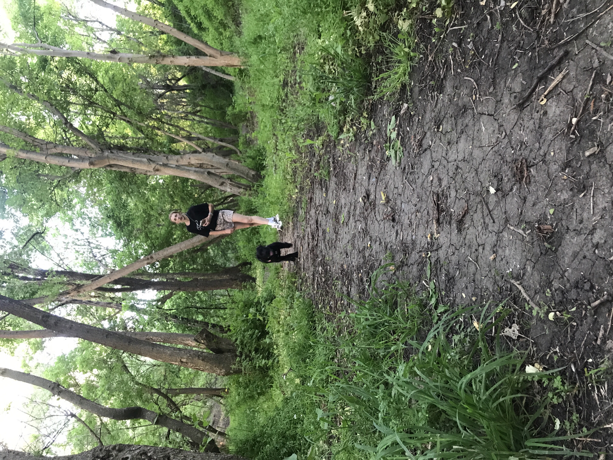 The image shows a woman standing on a narrow dirt path in a green area, possibly a park or forest. A black dog stands a few feet in front of her. The path is bordered by grass and dense vegetation. Trees line both sides of the path, creating a canopy overhead. The location is reportedly in Kharkiv, Ukraine.