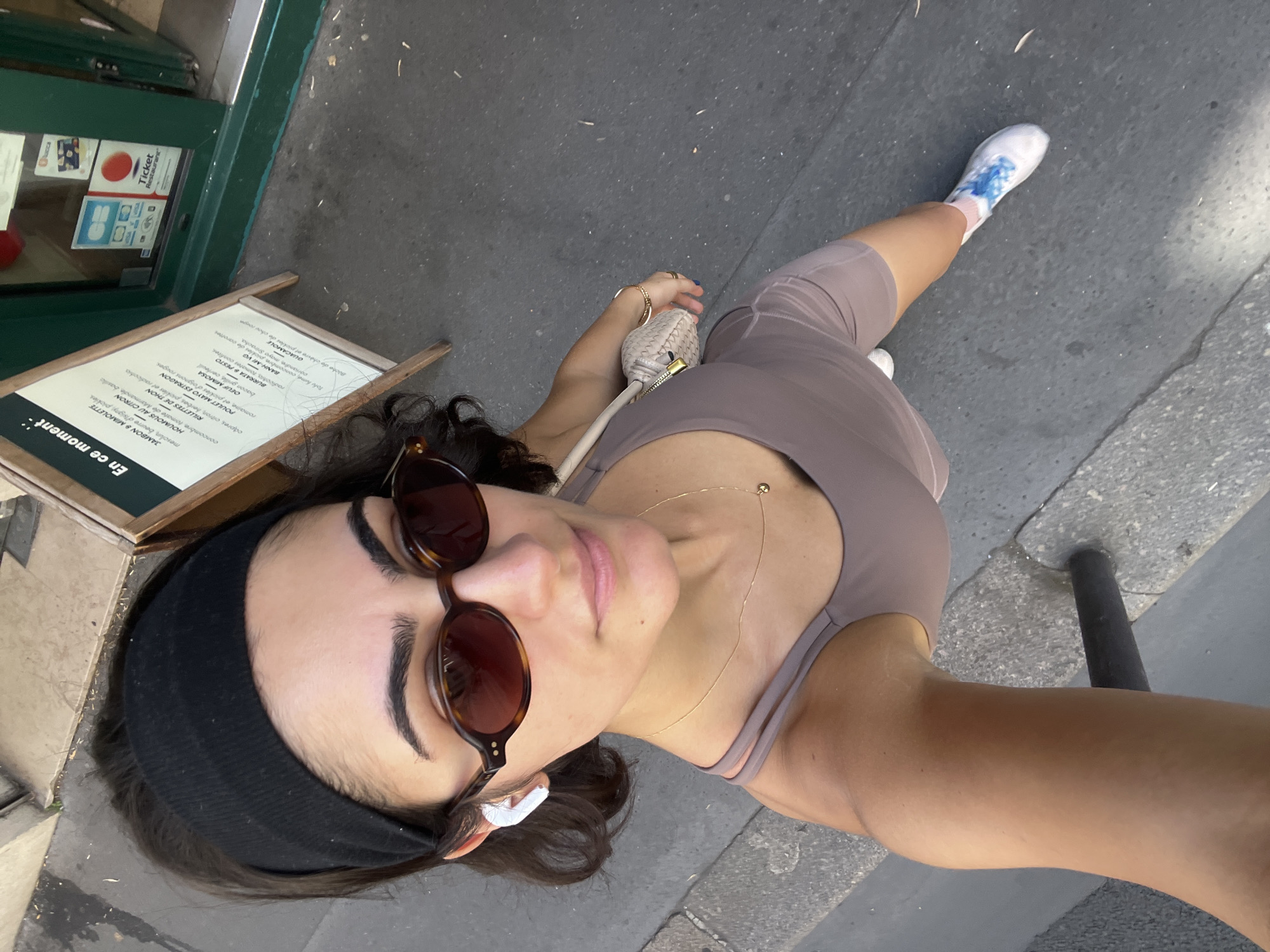 A woman with dark hair, wearing a black headband, sunglasses, a taupe tank top, and bike shorts, poses for a selfie on a street in Paris, France. She has white earbuds in her ears and a small, woven handbag slung over her shoulder. A wooden A-frame sign with a menu is visible to her right. The street is paved with asphalt, and a grey curb is on her left. She is wearing white sneakers with blue and pink accents. The overall scene is bright and outdoors.