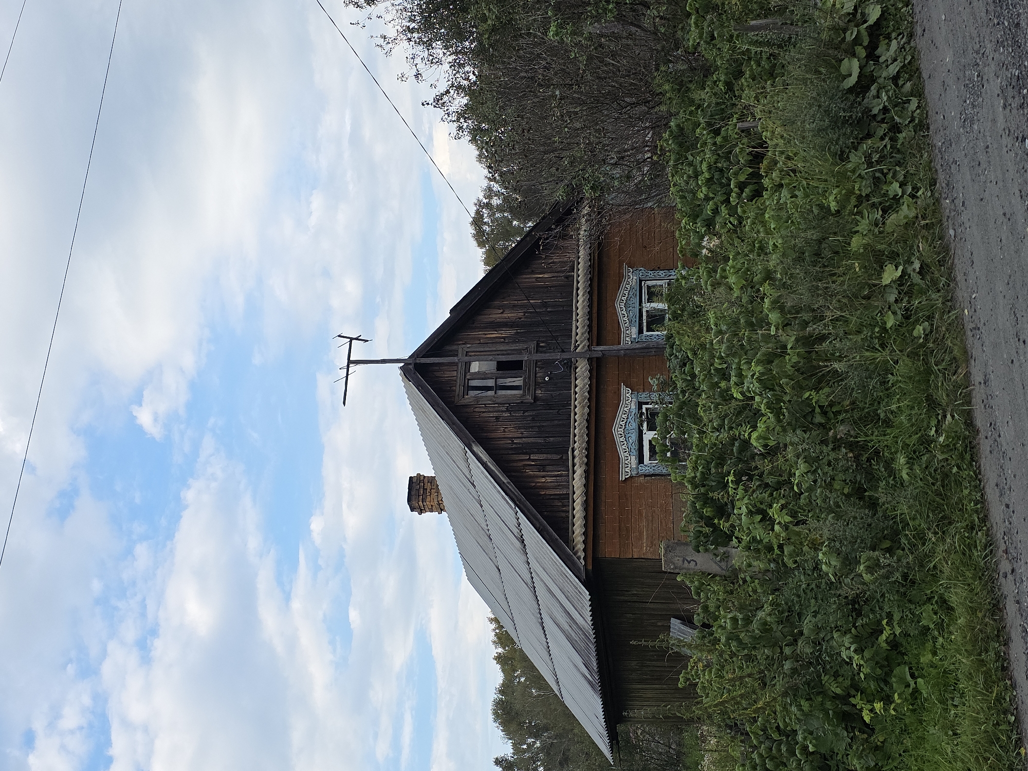 This is a full-shot, eye-level, exterior, natural light photograph of a dilapidated wooden house with a brick foundation in Magnitka, Russia. The house has a weathered, dark wooden upper story with a single window and a protruding chimney made of red brick. The lower story is constructed of orange brick and features two windows with intricate white and blue carved wooden frames. A corrugated metal roof extends over the house. The house is surrounded by overgrown green foliage, including bushes and tall grass, obscuring the lower half of the structure. A dirt road with gravel is visible in the foreground. A simple television antenna is mounted on a pole attached to the side of the house. The sky above is a mix of blue and white clouds. The scene appears to be located in a rural or semi-rural area, as suggested by the unkempt vegetation and the style of the house.