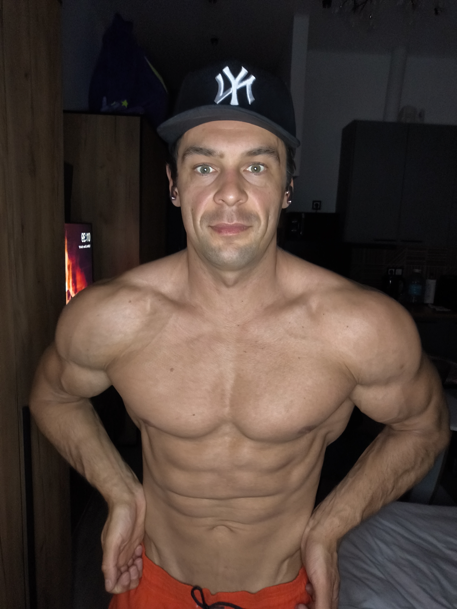 A shirtless man wearing a black "NY" baseball cap is posing with his hands on his waist, displaying his muscular physique. He appears to be in a dimly lit interior setting. A television is visible in the background to the left, displaying the time as 08:10, along with a burning image. Cabinets are visible behind the subject, possibly in a kitchen or living area. There is no evidence to suggest the location is in Moscow, Russia.