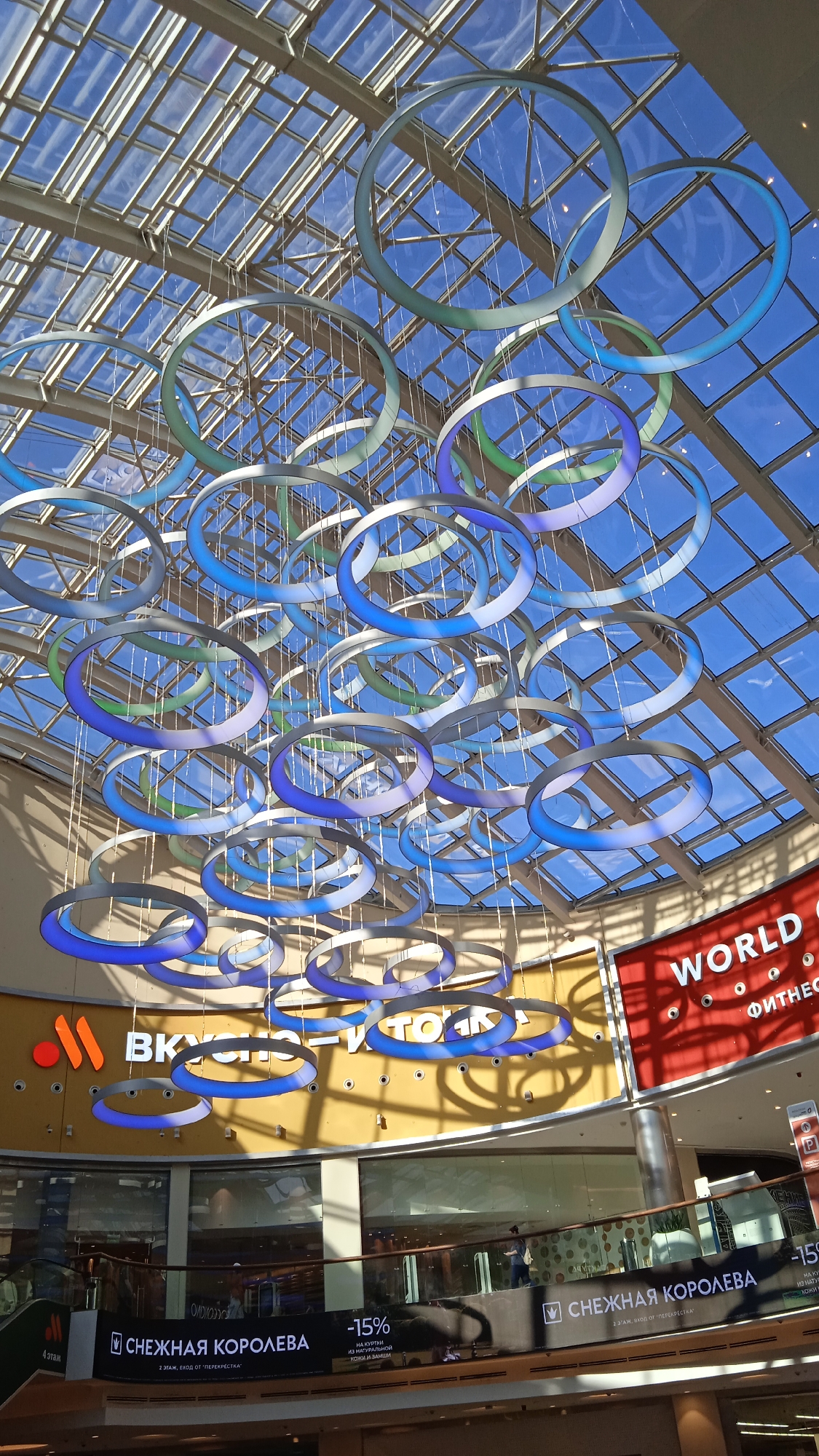 This is an indoor shot of a shopping mall in Khimki, Russia. The image focuses on a large, modern ceiling installation of interconnected rings, some illuminated with blue and green light. The ceiling itself is a glass and metal structure, allowing natural light to enter, creating a bright atmosphere. Below the rings, a large yellow wall features the orange and white logo of "ВКУСНО И ТОЧКА" (Vkusno i tochka), a fast-food chain that replaced McDonald's in Russia. To the right of this, a red sign displays "WORLD OF FITNESS" in white lettering. Further down, a store named "СНЕЖНАЯ КОРОЛЕВА" (Snezhnyy Korolev / Snow Queen) is visible with a banner advertising "-15%" discounts on natural fur coats and hats. A few shoppers are present on the lower levels, one walking on a balcony. The overall impression is of a contemporary, well-lit commercial space.