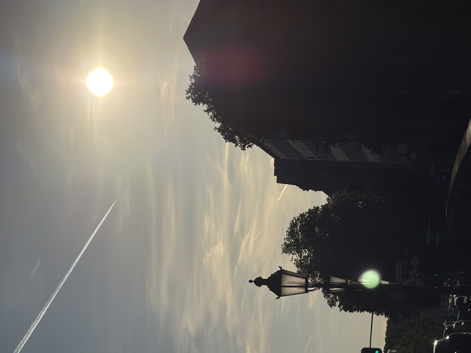 The image captures a street scene in Munich, Germany, under a bright, slightly hazy sky. A prominent jet contrail stretches diagonally across the upper left portion of the frame. The sun, appearing as a bright, diffused orb with a lens flare, is positioned in the upper right quadrant. In the foreground, a dark silhouette of an ornate street lamp stands to the left, its glass panels visible. To the right, the dark silhouettes of buildings and trees dominate the scene. One building shows a repetitive pattern of windows. The lower part of the image depicts a street with vehicles, including car headlights, and a traffic light displaying green. A green light also reflects on the street lamp. The overall impression is of a late afternoon or early evening urban environment.
