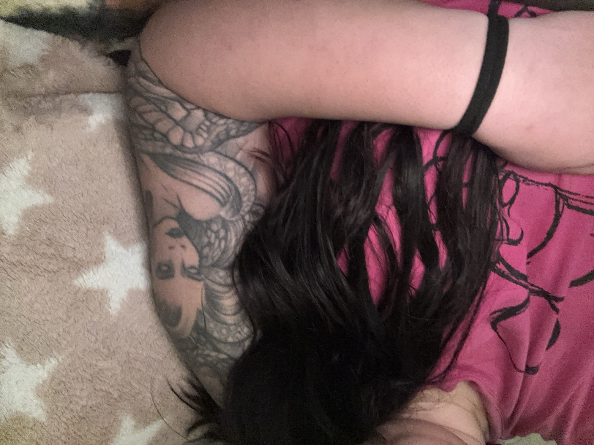A person is lying on a bed. The person is wearing a pink shirt with black abstract design. They have black hair that is partially covering the shirt and an arm. There is a black elastic band around their other arm. The arm that is visible has a detailed tattoo of a woman and a snake on it. The bed has a patterned blanket. There is no evidence to suggest the location is Dobrovnitsa, Bulgaria.