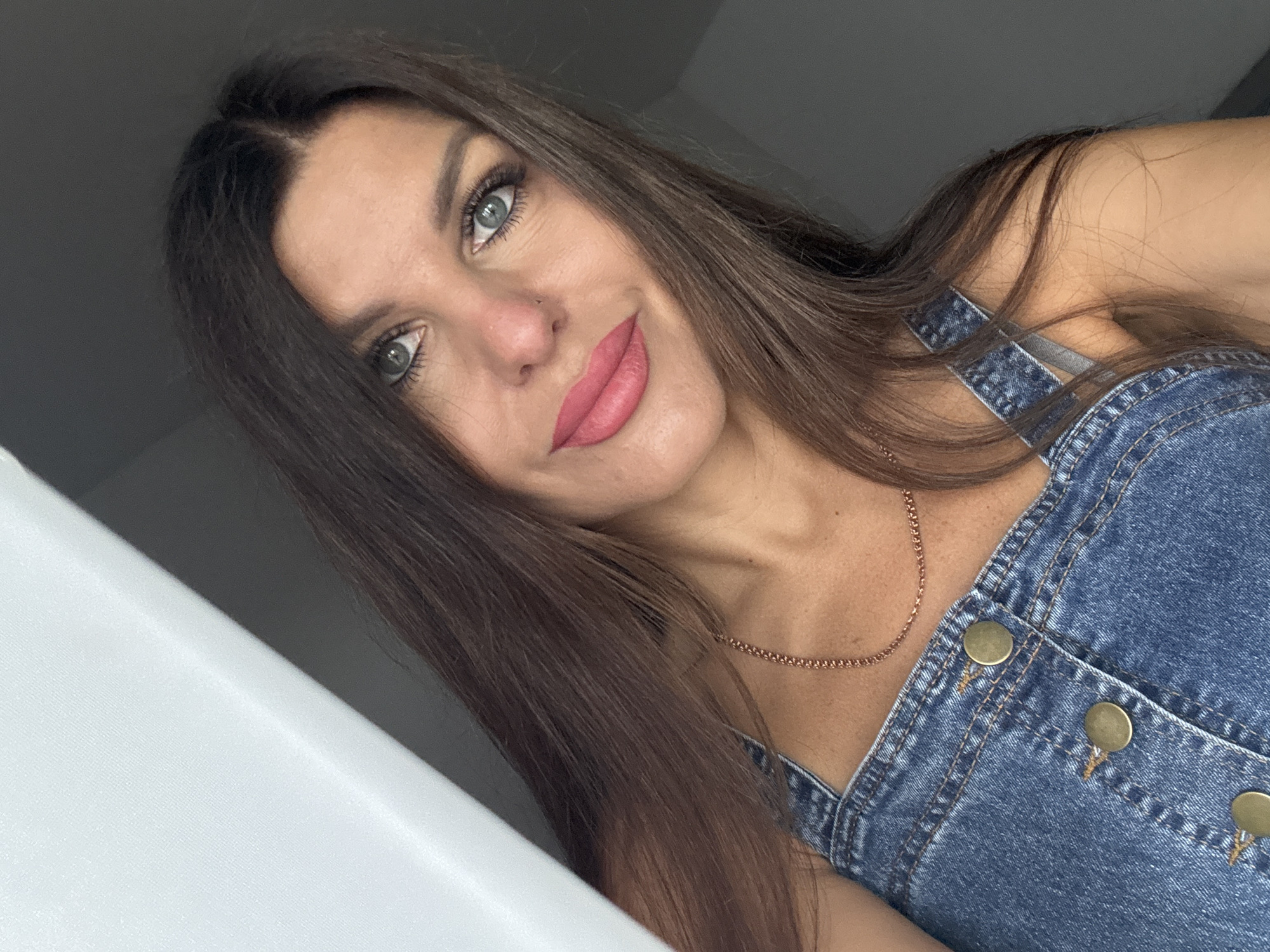 A medium close-up shot shows a woman with long, dark brown hair and green eyes. She is wearing a denim jumpsuit and a rose gold chain necklace. Her head is tilted to her left, and she is looking directly at the camera. Her lips are painted with a pink lipstick. The background is divided between a bright white surface on the left and a dark gray wall on the right. The lighting suggests an indoor setting, possibly near a window. No specific location in Chaikovsky, Russia, can be identified from the visual information.