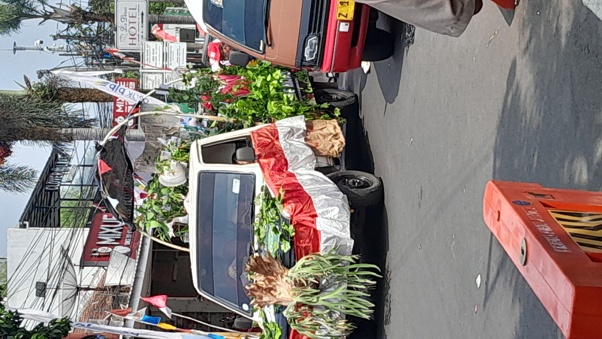 A white pick-up truck decorated with Indonesian flags and plants is parked on a street in Sumedang, Indonesia. The truck is adorned with a large staghorn fern on the hood, and the sides are draped with red and white fabric, resembling the Indonesian flag. The truck's bed is also filled with greenery and red flowers.
In the background, various buildings and businesses line the street, including one with a prominent "MIXUE" sign. Several flags and banners are visible, suggesting a festive or celebratory occasion. A brown minivan is parked to the right of the pick-up truck. The street surface is asphalt, and shadows suggest it is daytime. In the foreground, a red plastic traffic barrier is present.