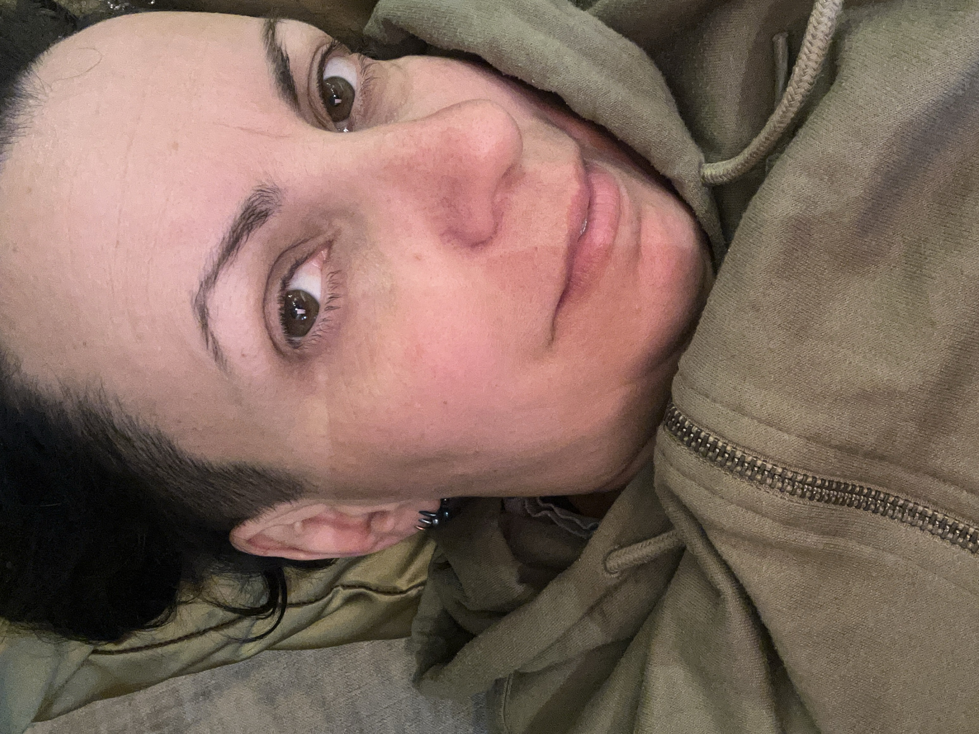 A close-up, slightly angled, selfie-style photograph shows a person with fair skin, brown eyes, and short, dark hair, wearing a green hooded sweatshirt. The person is lying down, with their head resting on a tan-colored surface with a grey texture, possibly a pillow or upholstery. The person has visible facial features including eyebrows, eyelashes, a nose, and lips. They are wearing a silver earring in their left ear. The hoodie's zipper is visible. There is no indication of the location being Sofia, Bulgaria.