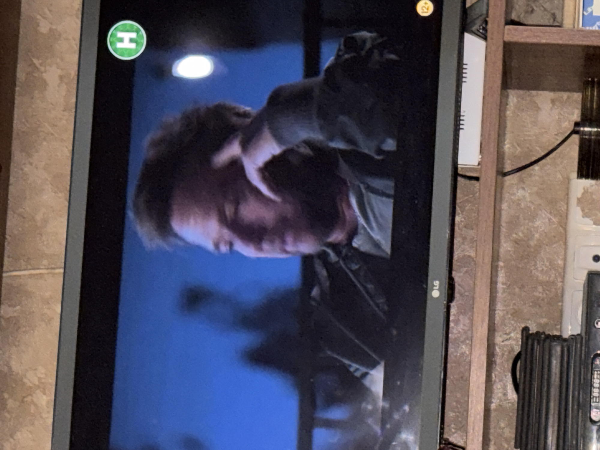 The image shows a television screen displaying a man's face from the shoulders up. The man has light-colored hair and is wearing a dark jacket over a lighter shirt. His left hand is resting on his chin, and his right hand is near his head. The background of the television screen is a dark blue with blurred figures and lights. In the top right corner of the screen, there is a green circular logo with the letter "H" inside. In the bottom right corner of the screen, there is a small yellow circle with "12+" inside. The television is placed on a wooden shelf, with a textured wall visible behind it. Below the television, there are some electronic devices and cables.