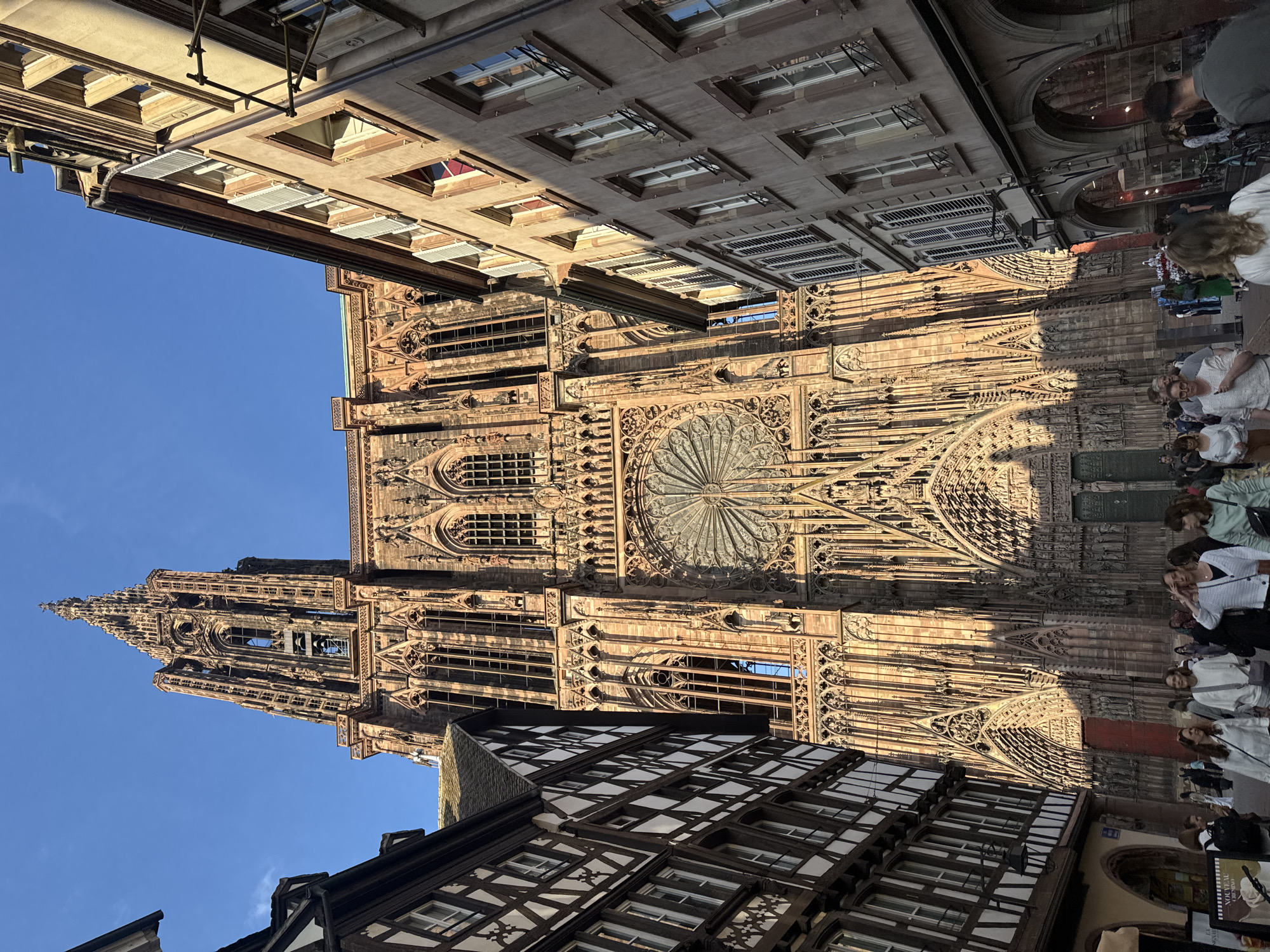 The image shows the Strasbourg Cathedral (Cathédrale Notre-Dame de Strasbourg), a significant landmark in Strasbourg, France. The cathedral's west facade, with its intricate stonework, rose window, and towering spire, dominates the view. The sky is clear and blue. At the base of the cathedral, a crowd of people are seen. Traditional timber-framed buildings typical of the Alsace region frame the cathedral. A scaffolding system is partly visible at the top.
