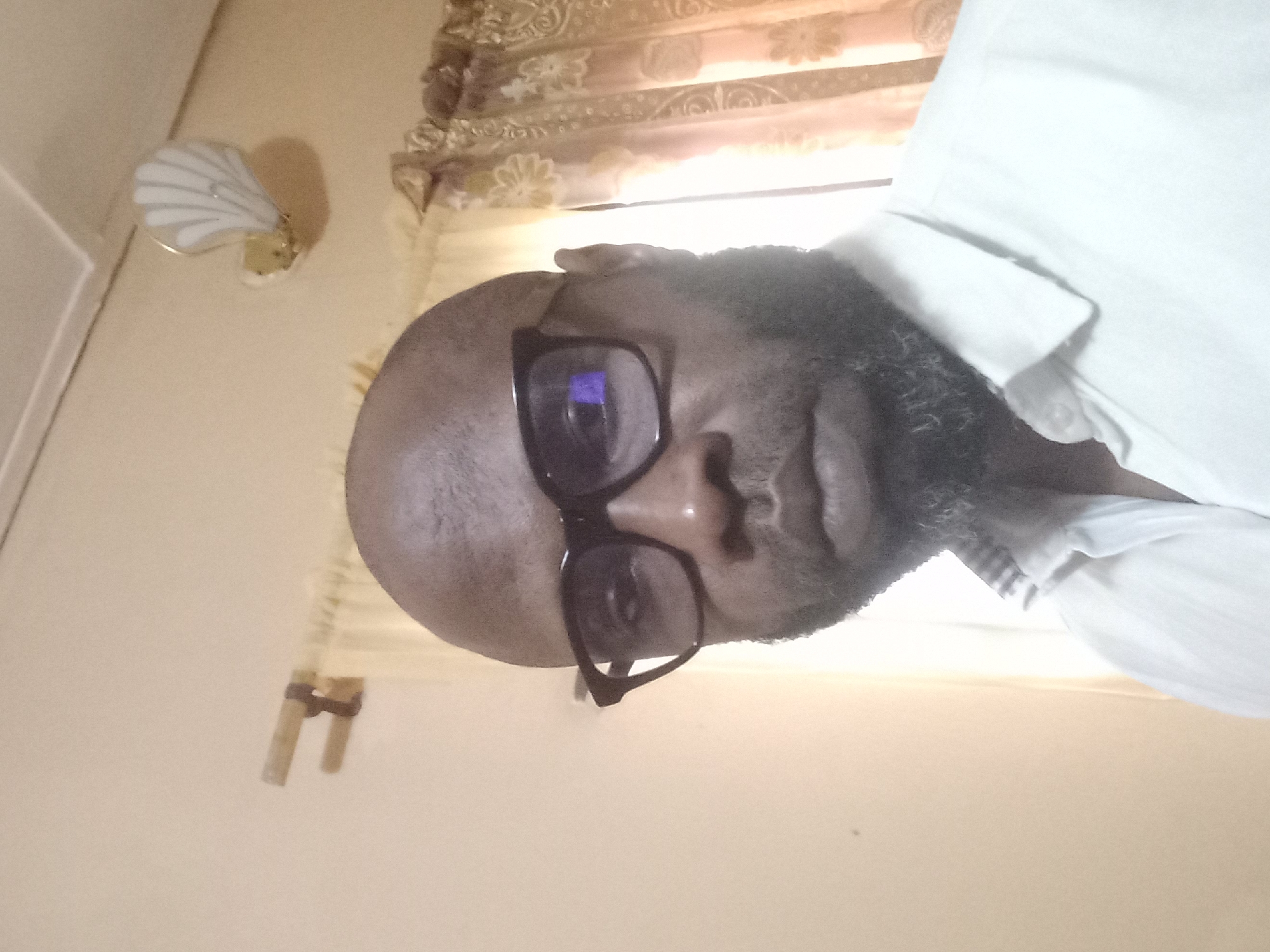A close-up shot depicts a Black male wearing glasses and a white collared shirt. He is positioned against a background with beige walls and patterned curtains. An object resembling a wall sconce is visible on the wall behind him. The location is Barden Yana, Nigeria.