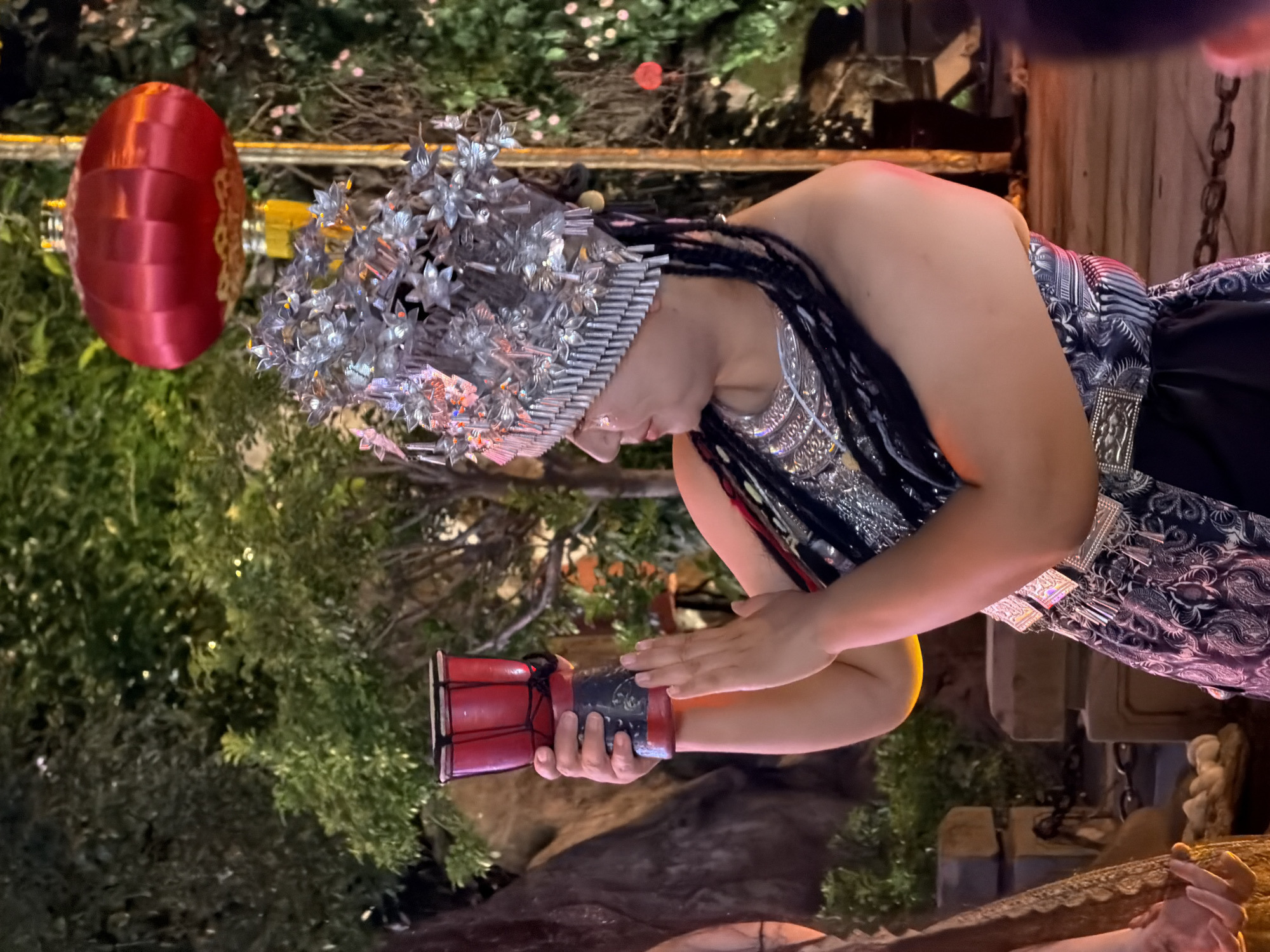 A person dressed in traditional attire is holding a small red drum and performing in what appears to be an outdoor event in Yuzhong District, China. The person wears an elaborate silver headdress adorned with floral motifs and numerous hanging ornaments. They are also wearing a beaded necklace and a patterned garment with silver embellishments. The background features lush green foliage, a bright red lantern hanging from a pole, and stone structures. The lighting suggests it is evening or nighttime, with ambient lights illuminating the scene. The person's hands are positioned as if playing or about to play the drum. The overall atmosphere seems festive and cultural.