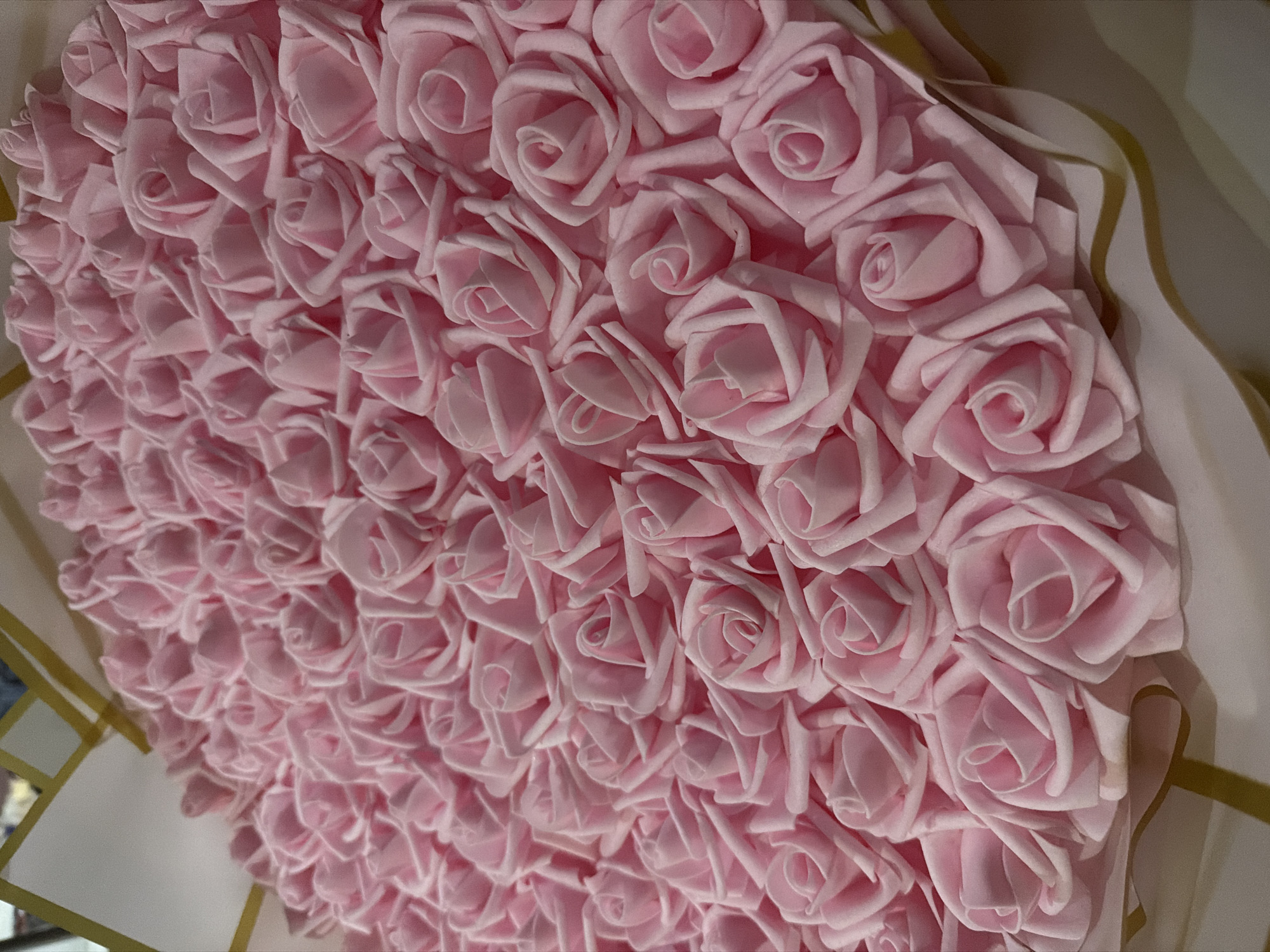 The image displays a close-up, angled view of a dense arrangement of numerous light pink artificial roses. The roses, likely made of foam or fabric, are uniformly shaped with tightly spiraled centers, creating a textured surface. They are packed closely together, filling the majority of the frame.
The roses appear to be part of a larger decorative item, possibly a large bouquet, a heart-shaped display, or a gift arrangement. Along the right and bottom edges of the main image, there are hints of a lighter, possibly off-white or cream-colored, wrapping or backing material, accented with thin, wavy golden lines or ribbons. This suggests the roses are presented within a decorative context.
There is no activity taking place, as the image captures a still life. The soft pink color of the roses typically symbolizes gentleness, admiration, joy, and love, suggesting the item is a gift or decoration for a special occasion such as a birthday, anniversary, or Valentine's Day.
The lighting is soft and even, highlighting the contours of the individual rose petals and the slight shadows cast by each flower, adding depth to the composition. The time of day and weather are indeterminate, suggesting an indoor setting. There are no specific visual cues within the image itself to indicate the location as Cherni Vrah, Bulgaria. No visible text is present in the image.