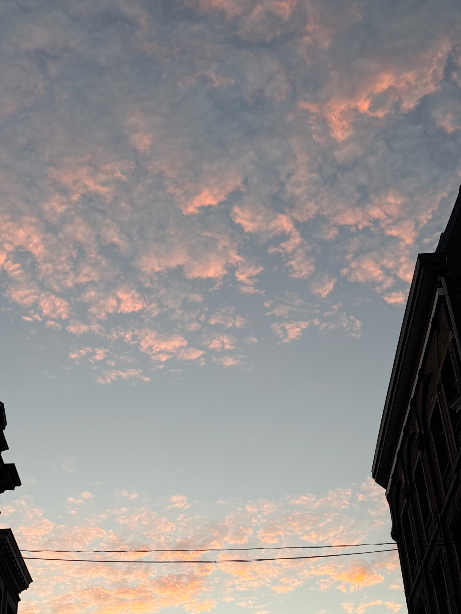 A view from below looking up at a sky filled with small, pink-tinged clouds. Two buildings, silhouetted against the sky, frame the scene on the left and right sides. There are power lines running horizontally across the lower portion of the image, between the buildings and the sky. The buildings appear to be made of brick and have arched windows. The overall impression is that of a sunset or sunrise over an urban environment, possibly in Liège, Belgium. There are no people or animals visible in the image.