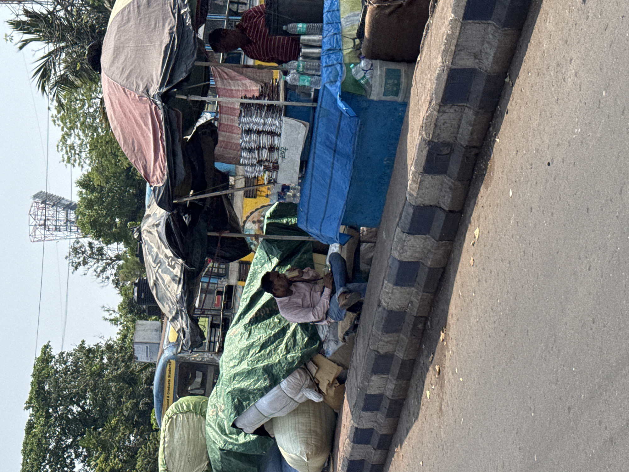 A low-angle, off-kilter shot in Kolkata, India, depicts a street scene adjacent to a pedestrian sidewalk. At the bottom of the frame, a segment of asphalt road is visible, meeting the raised sidewalk edged by black-and-gray checkered concrete blocks. An individual is reclining under a makeshift shelter constructed from various materials, including green tarps draped over sacks and merchandise. Further back, a vendor stands near a food stall displaying goods under awnings. The scene is framed by lush green trees and a distant metal structure against a clear sky.