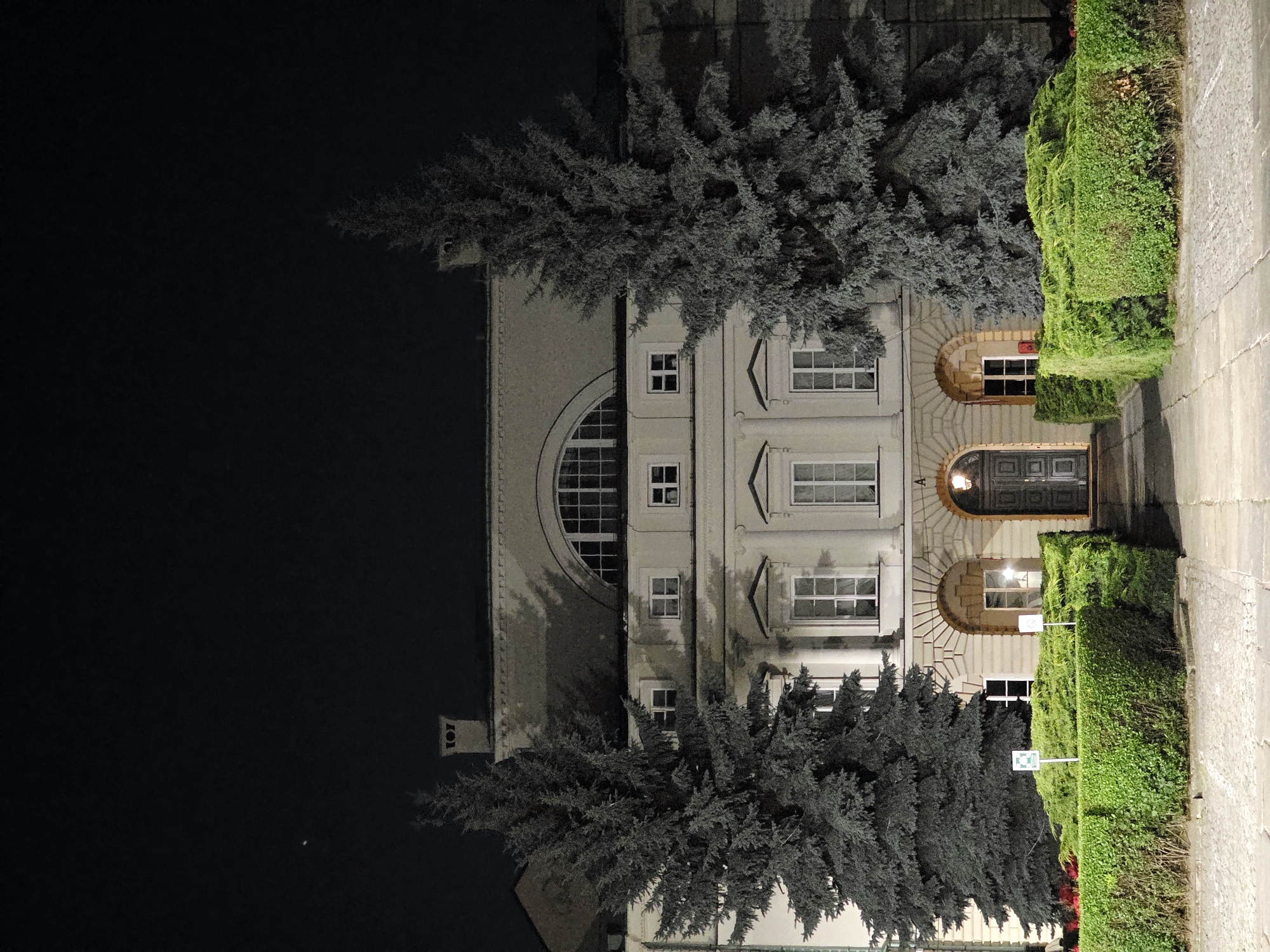 The image shows a building facade at night. The building is light-colored with multiple windows and an arched entranceway. There are several conifer trees with greyish-blue foliage in front of the building. To the right of the building, there is a neatly trimmed green hedge and a paved walkway. The scene is illuminated by artificial lights, casting shadows and highlighting the architectural details. A street number "107" is visible on a rectangular plaque on the left side of the building. There are also two white signs with green borders and symbols within the hedge. The overall impression is one of a well-maintained building and its surroundings, presented in a somewhat dramatic, nighttime setting.