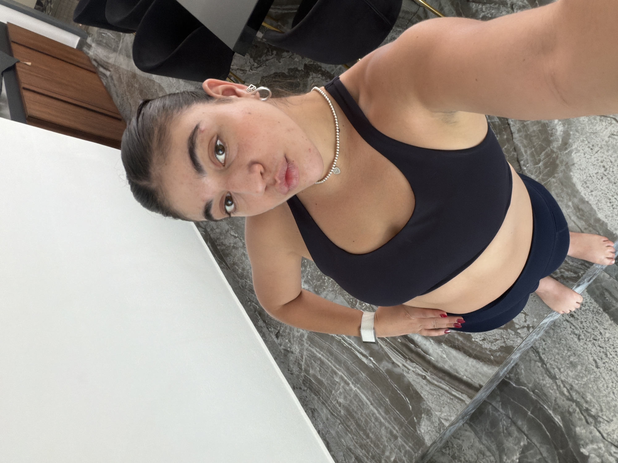 A young woman takes a high-angle, slightly tilted selfie indoors in Puebla, Mexico. She looks directly at the camera with a neutral expression and a subtle pout. Her dark hair is pulled back, and she wears a silver ear cuff on her left ear. She is dressed in a black sports bra and dark athletic leggings.
Around her neck, she has a silver beaded necklace with a small heart-shaped pendant, which may bear branding like Tiffany & Co. A white smartwatch or fitness tracker is visible on her left wrist, and her fingernails are painted a deep red. Her bare feet are visible in the lower right, resting on the floor.
The indoor setting features a polished, grey and white marbled floor. A plain white surface, possibly a wall or large piece of furniture, is prominent on the left side of the frame. In the background, dark, modern chairs and a wooden cabinet are partially visible. The lighting suggests it is daytime.